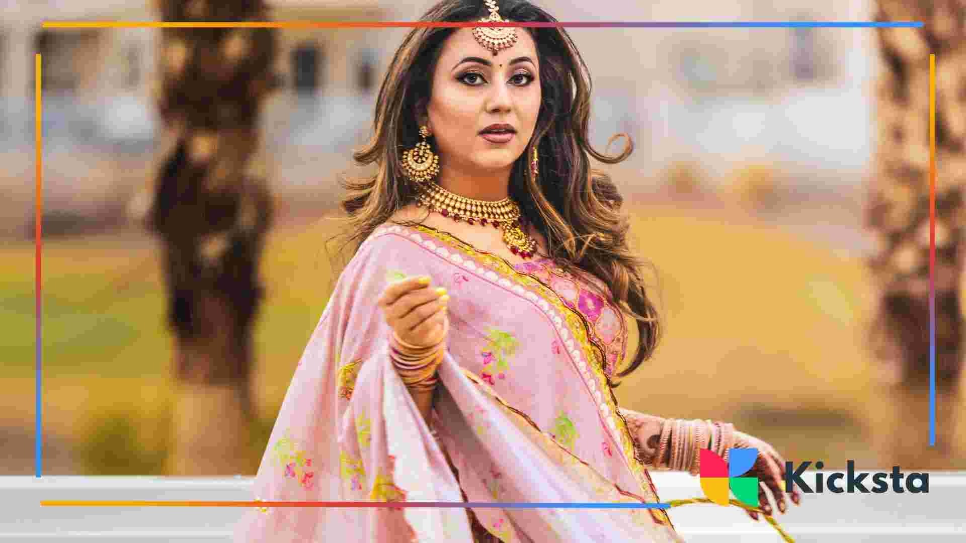 A woman dressed in a pink floral saree with gold jewelry poses outdoors, holding part of her saree.