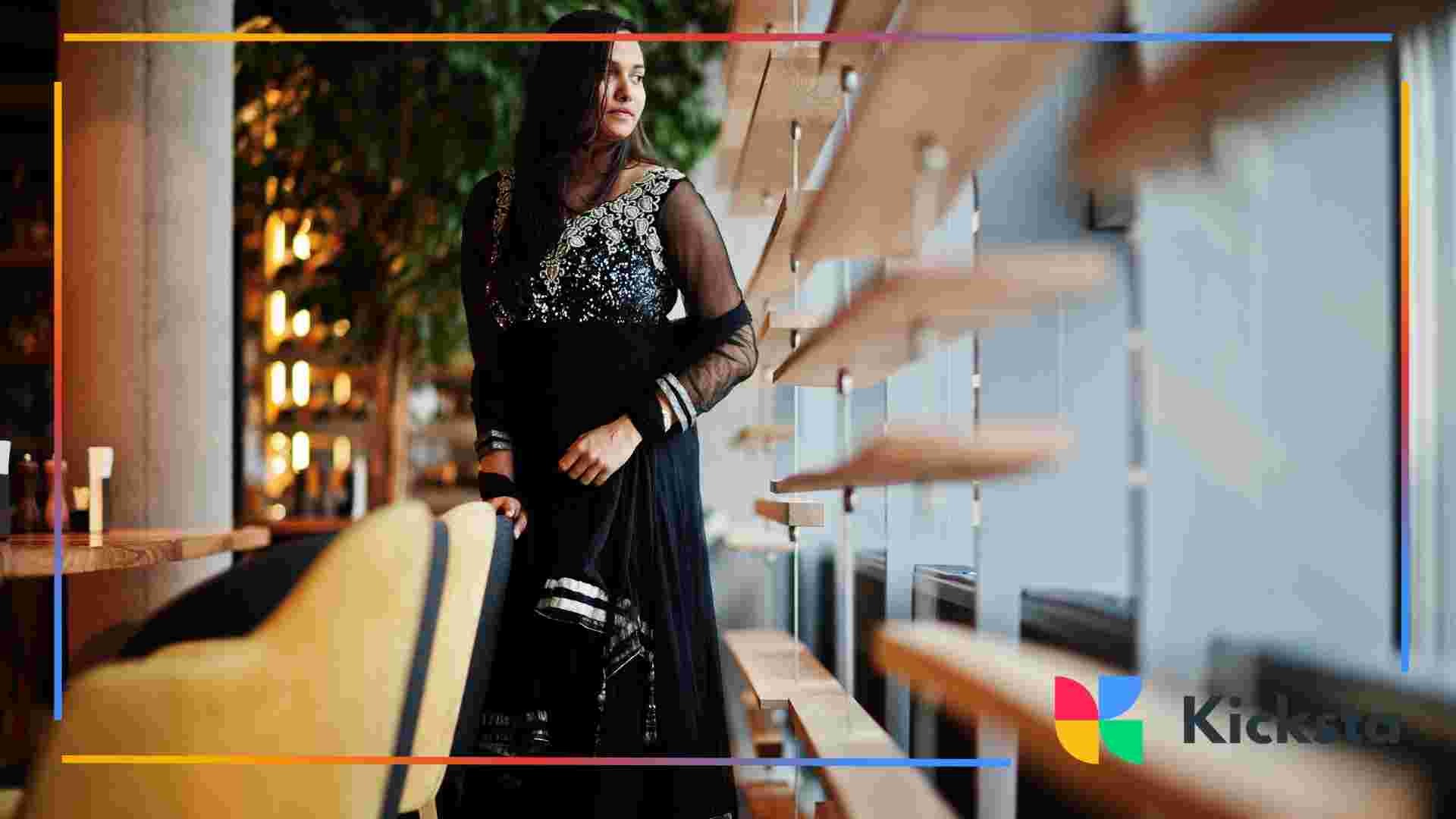 A woman wearing a black embroidered outfit stands by a window in a modern café, looking outside thoughtfully.