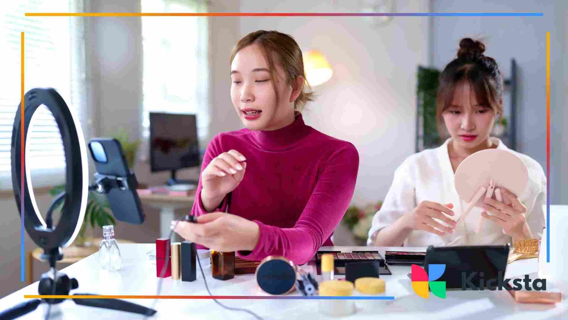 Two women sitting at a table creating beauty content, with makeup products, a ring light, and a phone set up for recording.