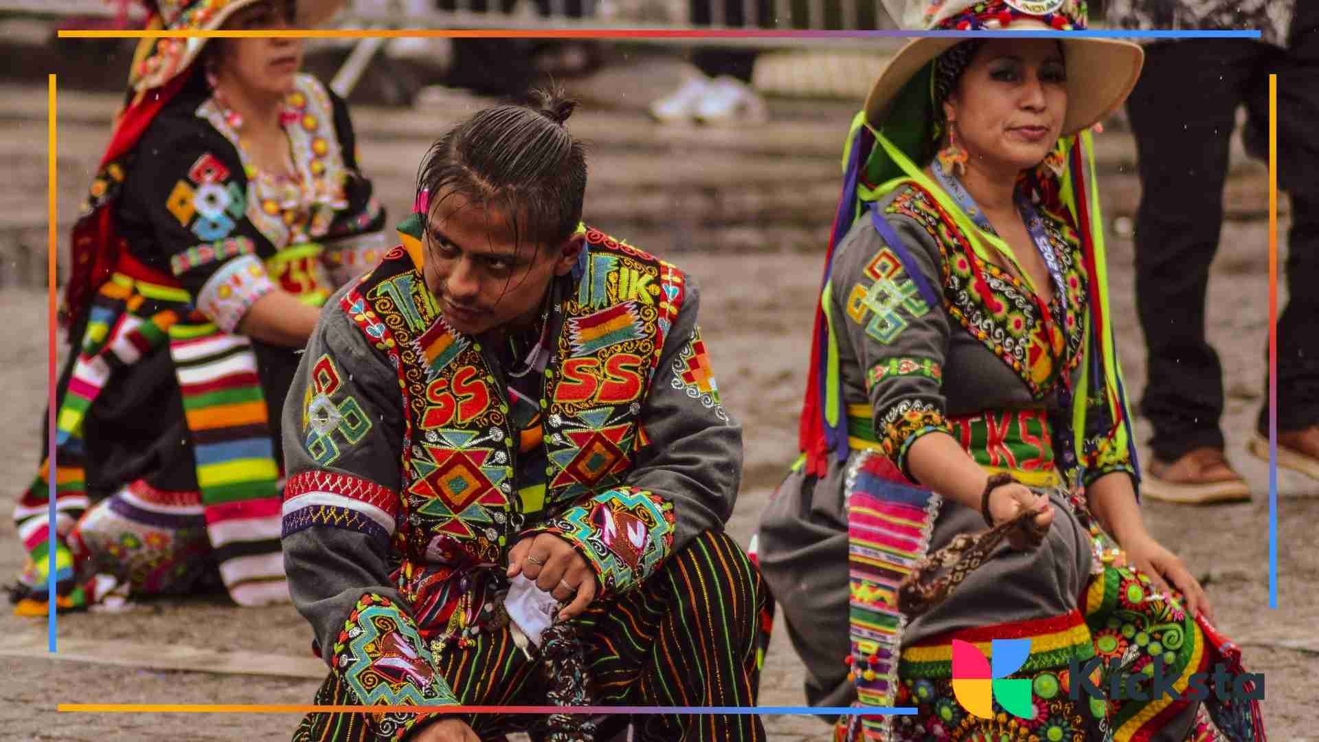 People dressed in vibrant Andean traditional clothing with multicolored embroidered patterns, sitting on the ground during a cultural performance or festival.