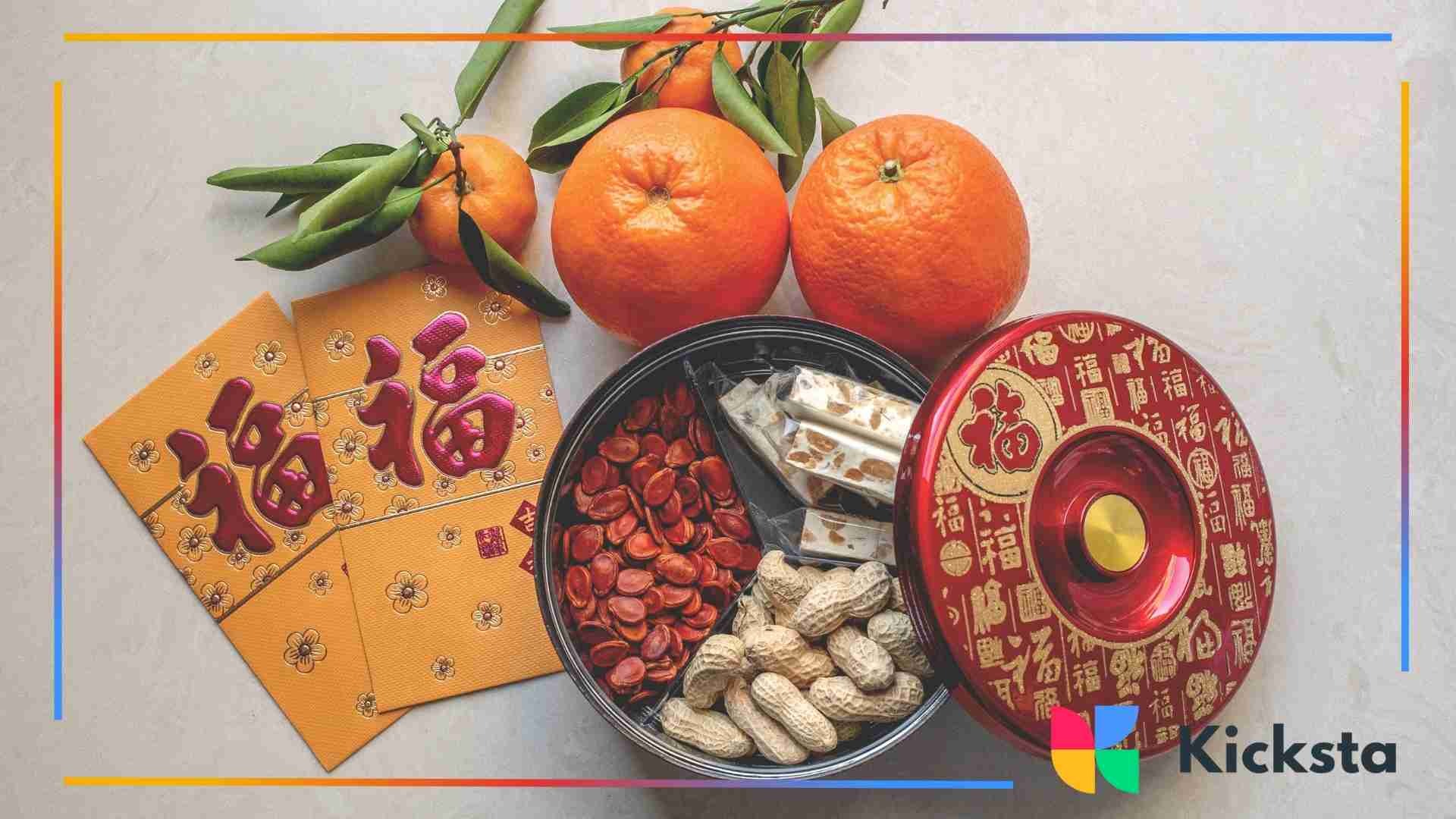 A flat lay of traditional Chinese New Year items including oranges with leaves, red envelopes with gold designs, and a red decorative container filled with peanuts, melon seeds, and candies.