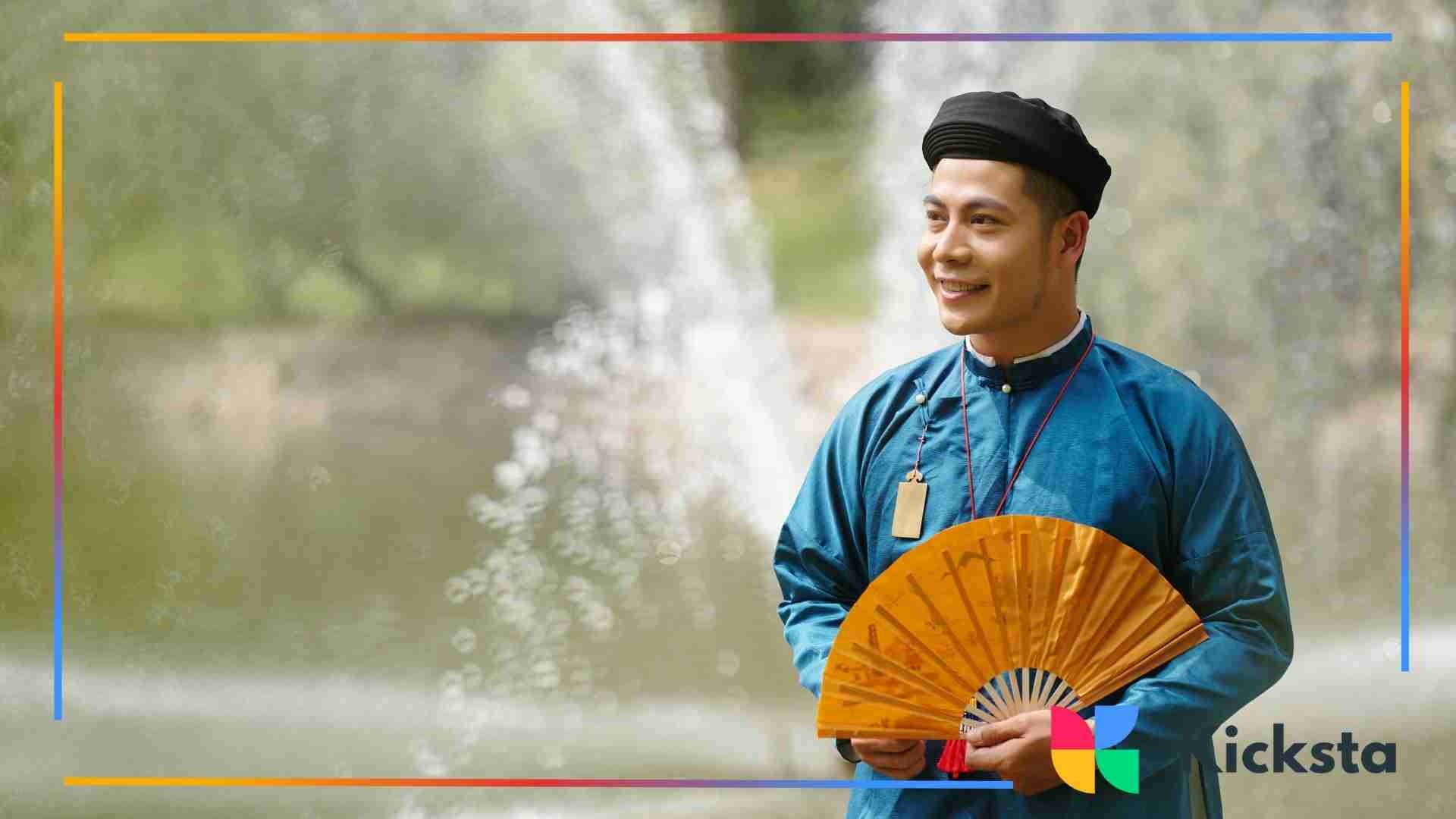 A man dressed in traditional Vietnamese clothing holding an ornamental fan, smiling while standing near a water fountain in a park.