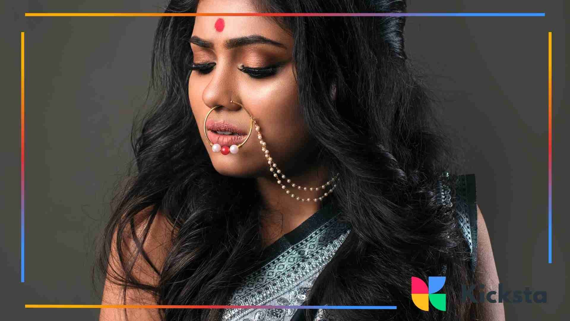 A woman wearing traditional South Asian attire and intricate jewelry, featuring a large nose ring connected to a chain of pearls, looking down with soft dramatic makeup.