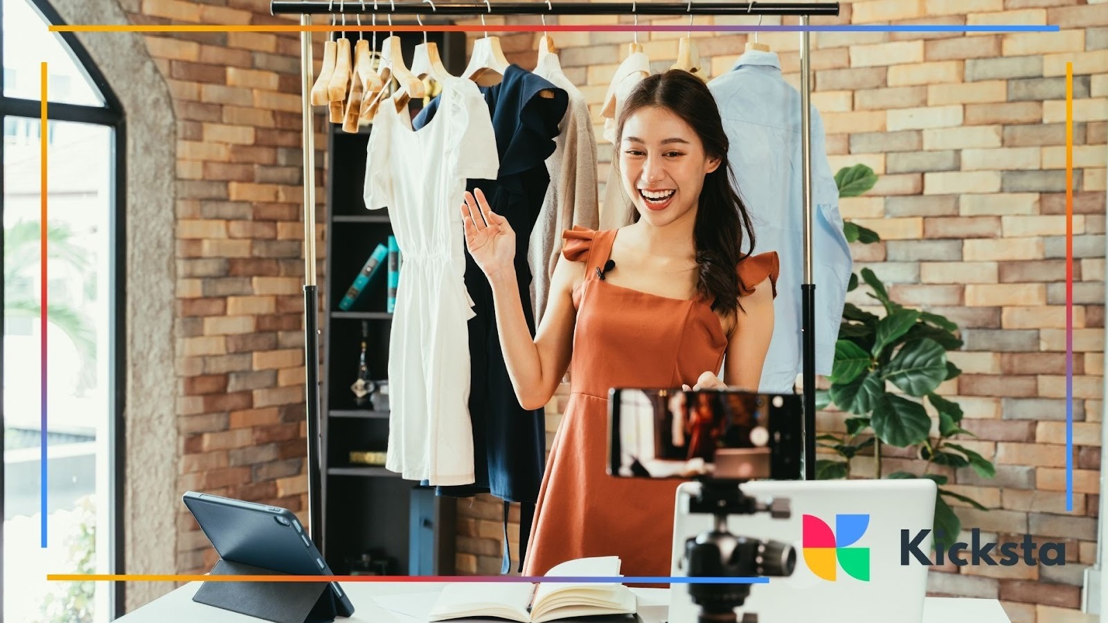 Woman hosting a live video in a boutique-style room, standing in front of a clothing rack and recording with a smartphone on a tripod while smiling and waving.