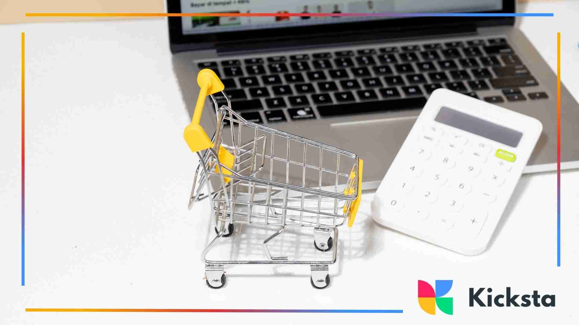 Small metal shopping cart and white calculator placed on a desk in front of an open laptop, symbolizing online shopping.