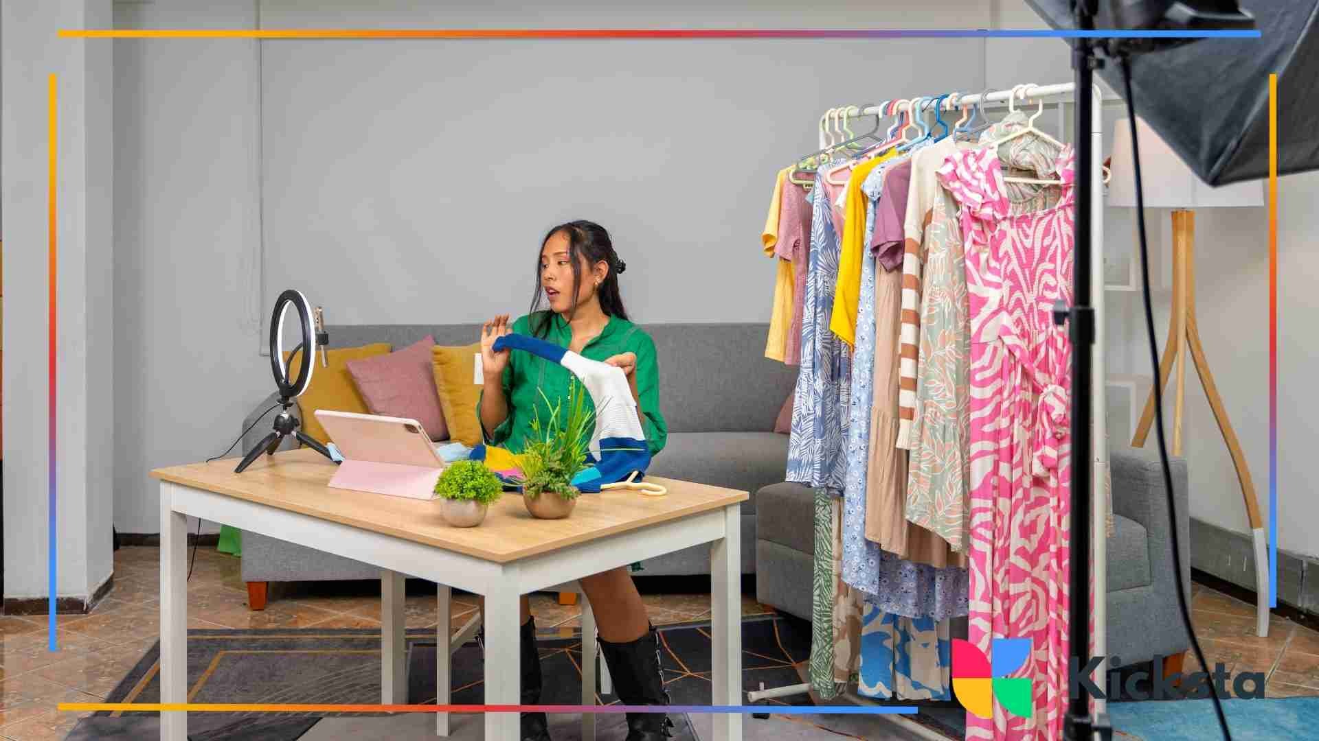 Woman recording a live selling video at home, holding clothing items while sitting at a desk surrounded by colorful garments and filming equipment.