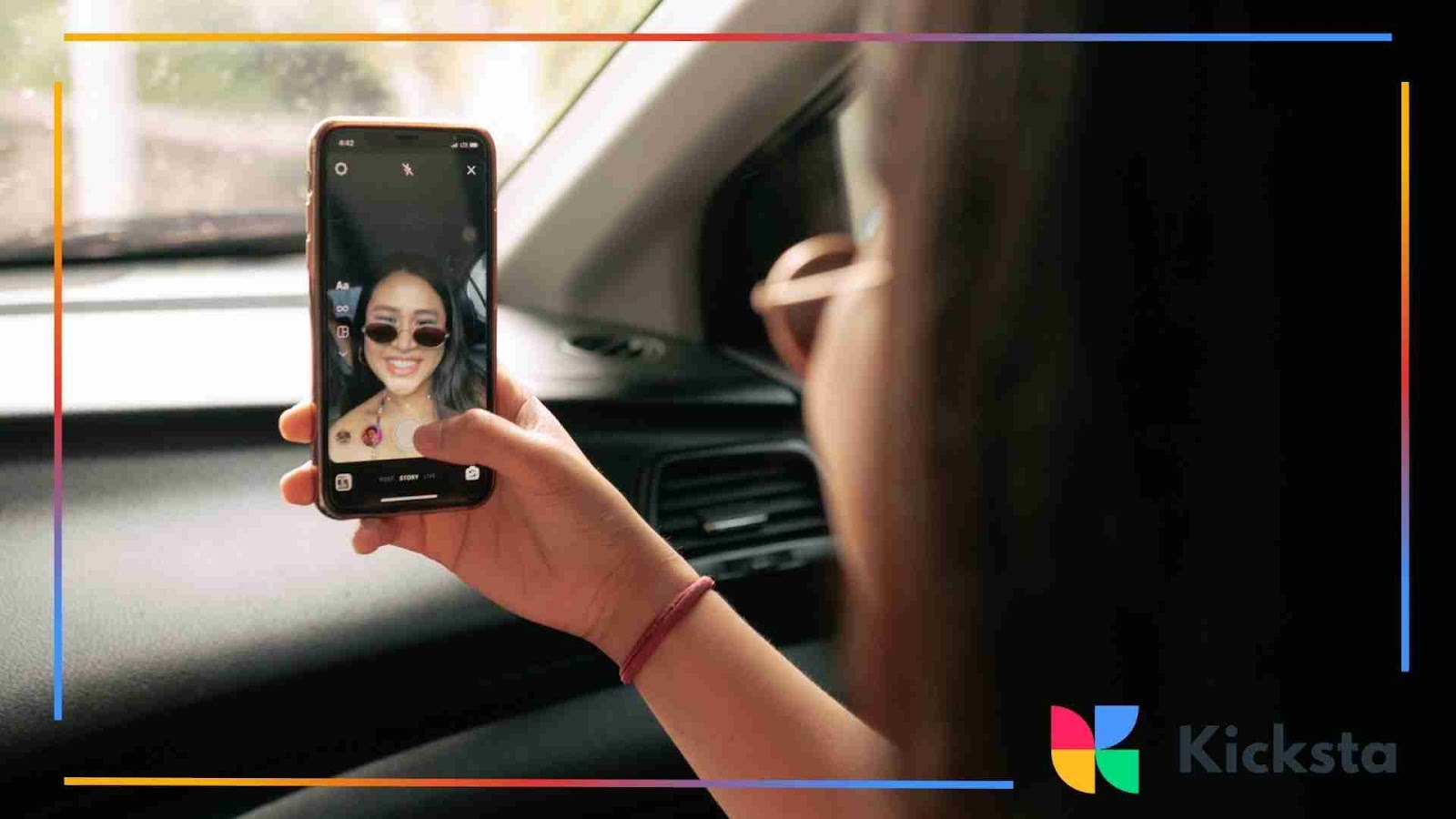A woman sitting in a car taking a selfie video on her smartphone, smiling while wearing sunglasses.