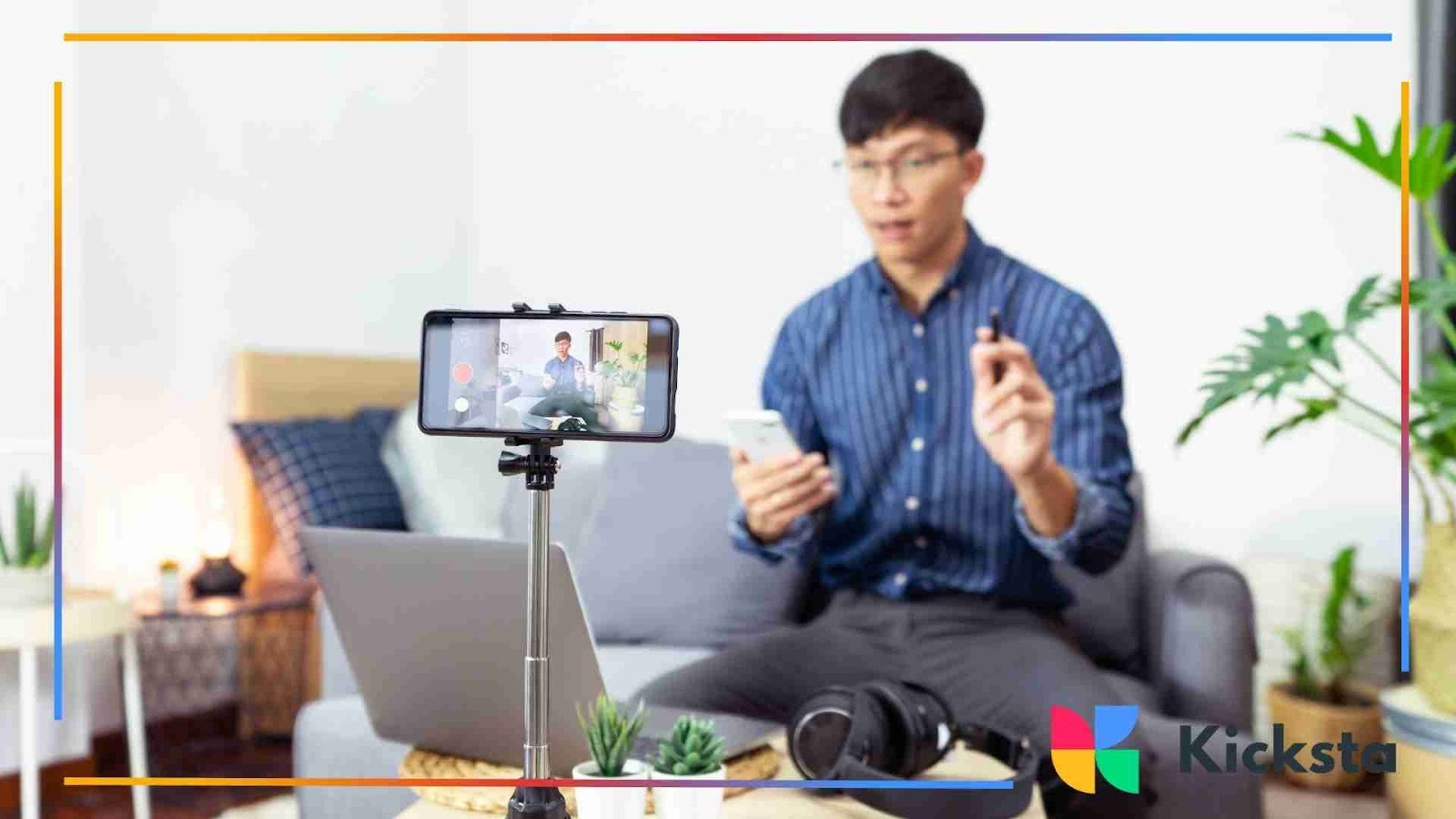 A man sitting on a couch filming content with a smartphone on a tripod, holding his phone and gesturing as he speaks.