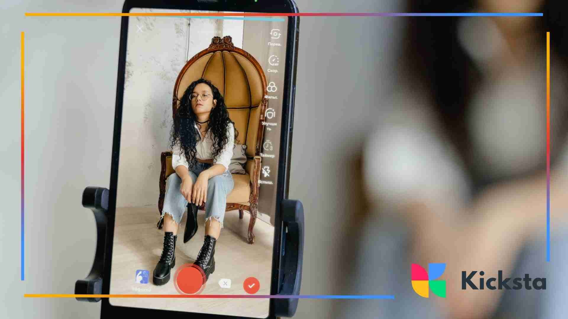 A smartphone on a tripod records a young woman with long curly hair sitting on a vintage high-back chair, posing for a video.