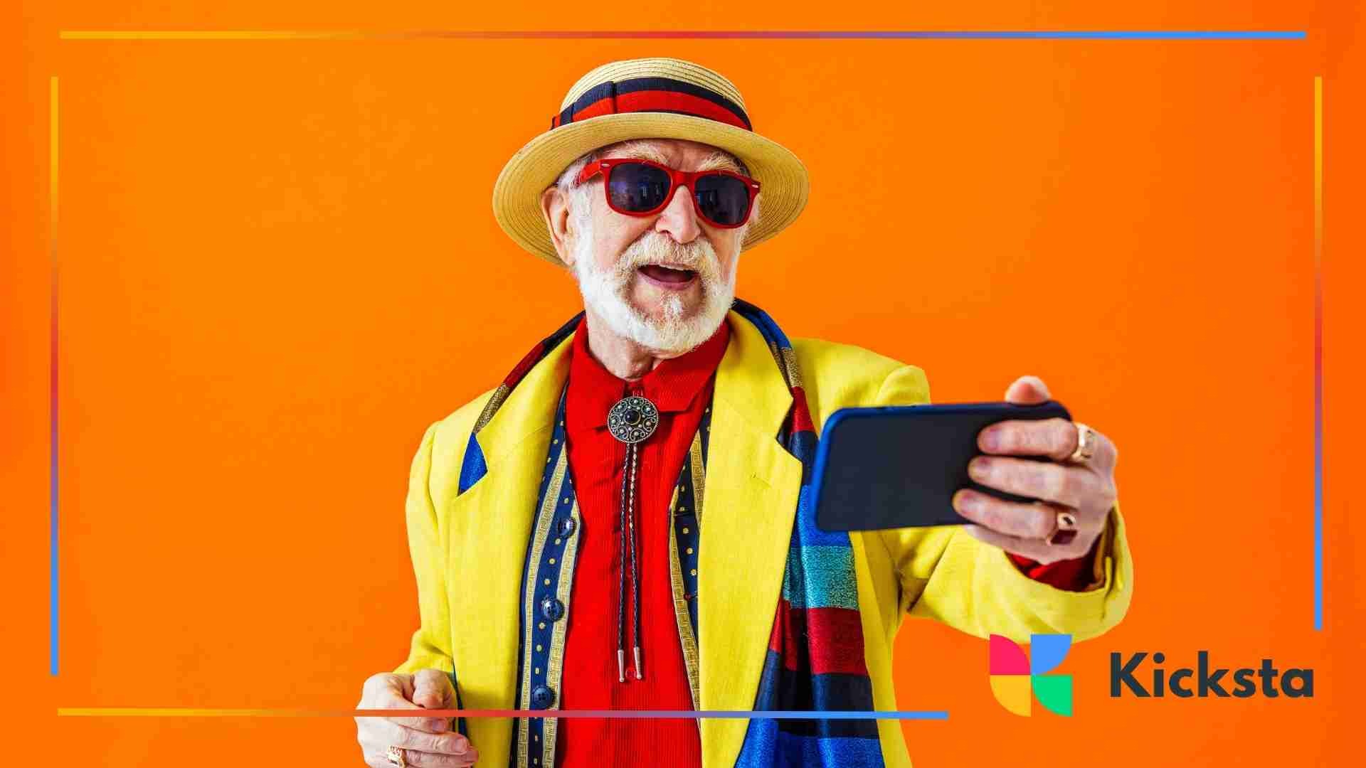 Stylish older man wearing bright yellow and red clothing, sunglasses, and a hat, holding a smartphone and recording himself against an orange background.