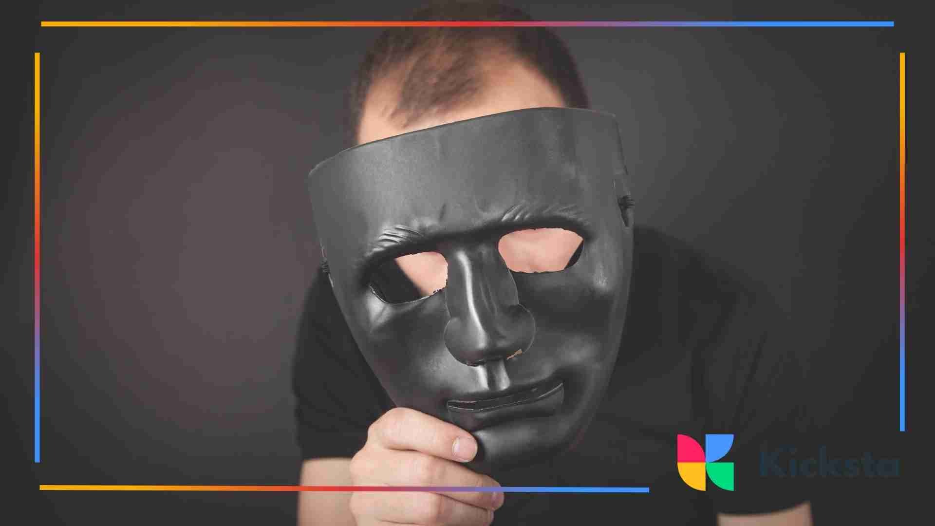 Person holding a large black mask in front of their face, covering most of their features. The background is dark.