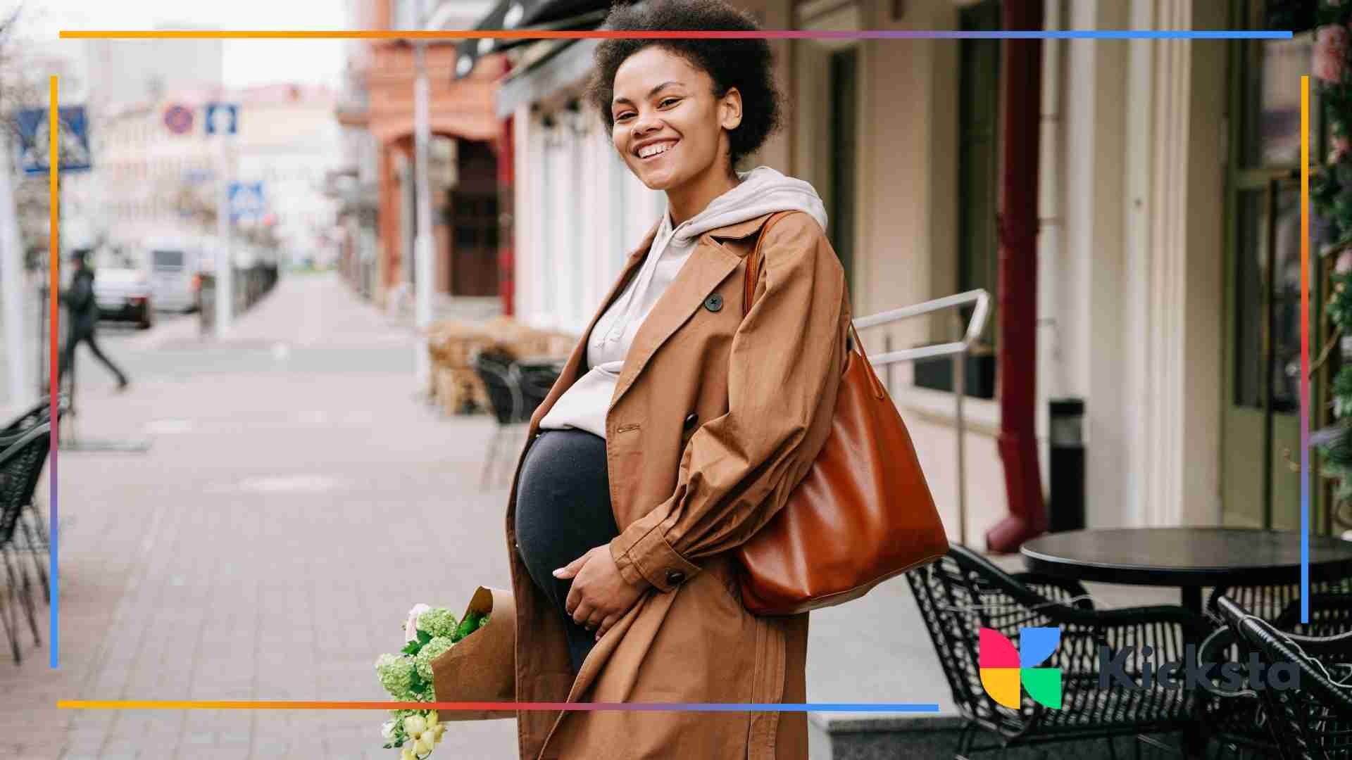A pregnant woman in a tan trench coat smiling while holding flowers and walking down a city street.