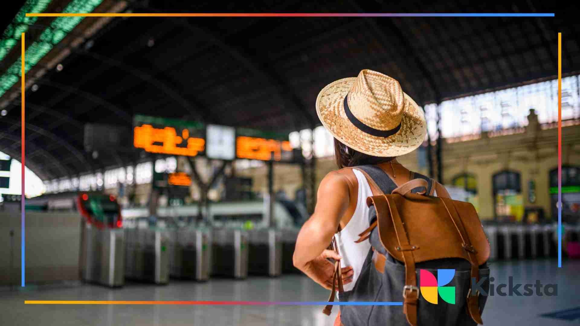 A traveler wearing a straw hat and backpack standing in a large train station, looking toward the departure signs.