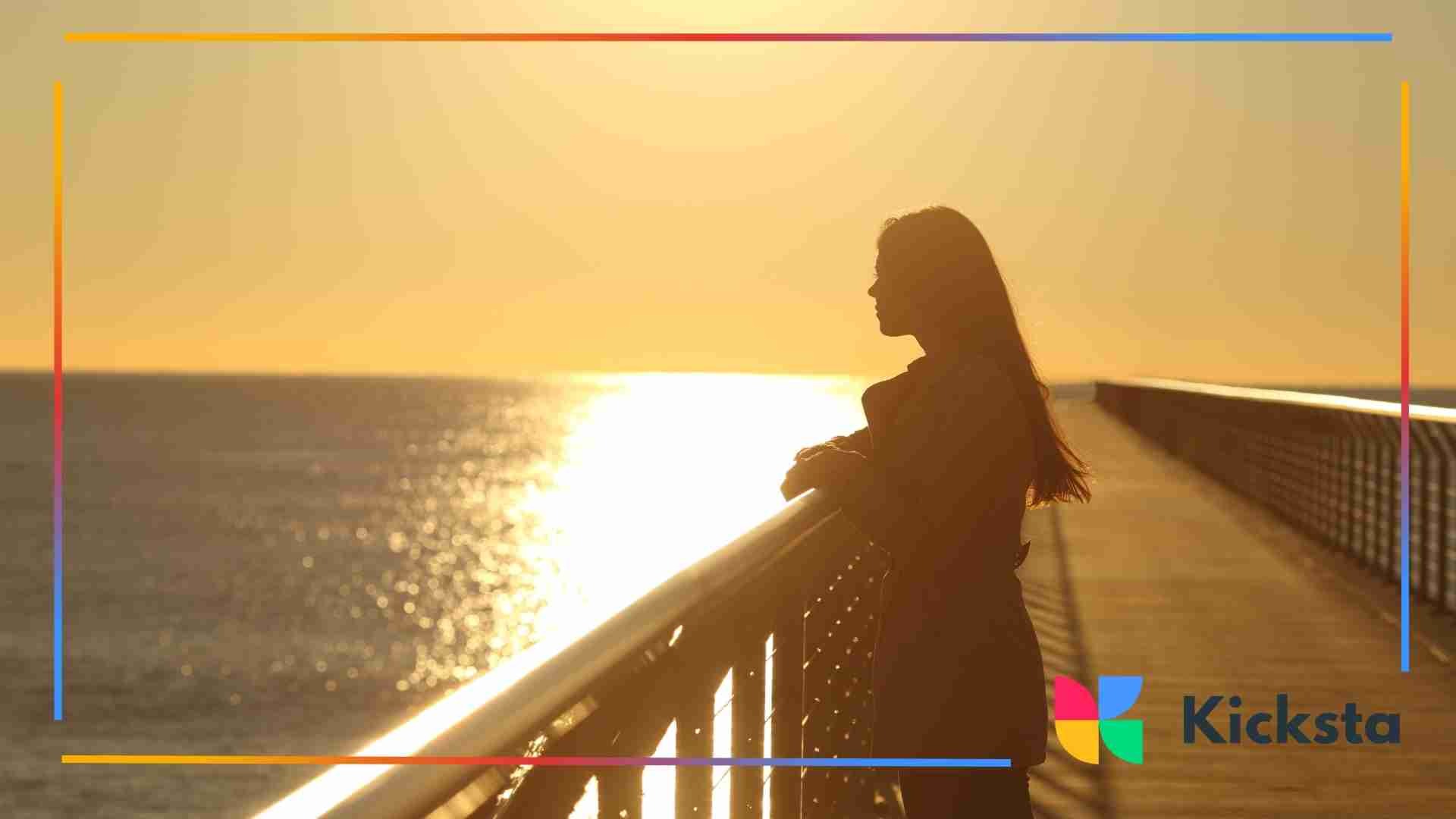Silhouette of a woman leaning on a railing while watching the sun set over the ocean.