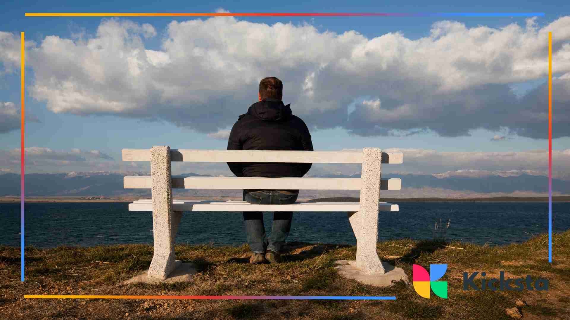 Man sitting alone on a white bench overlooking a large body of water with mountains in the distance.