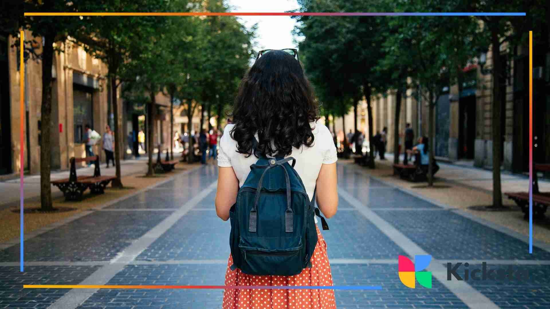 Back view of a woman with curly hair wearing a backpack while walking down a tree-lined city street.