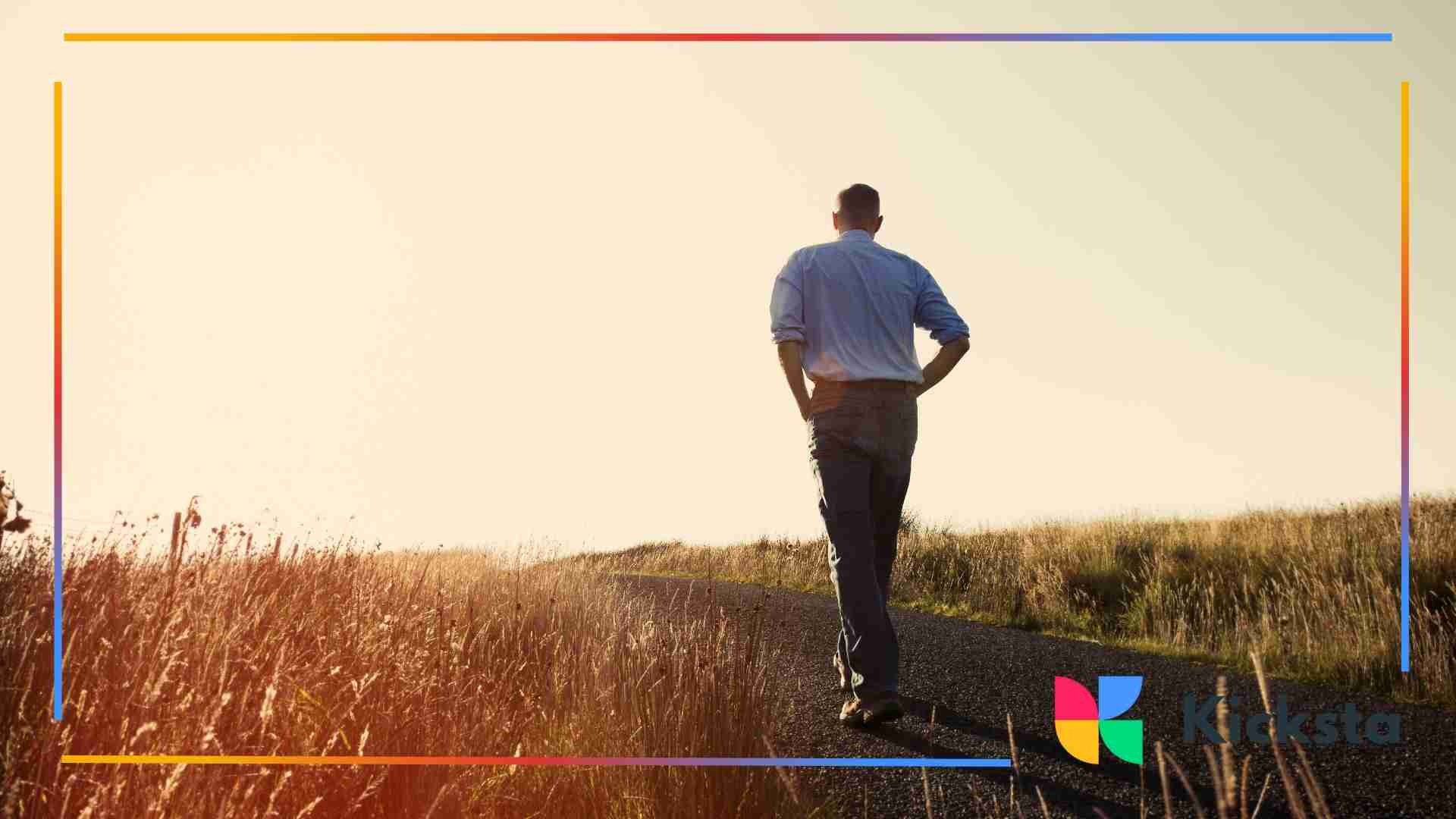 Man walking alone on a rural path surrounded by tall grass during golden hour.