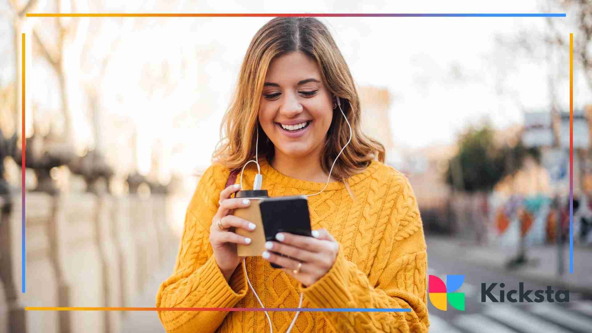 A woman in a yellow sweater smiles while looking at her phone and holding a coffee cup outdoors.