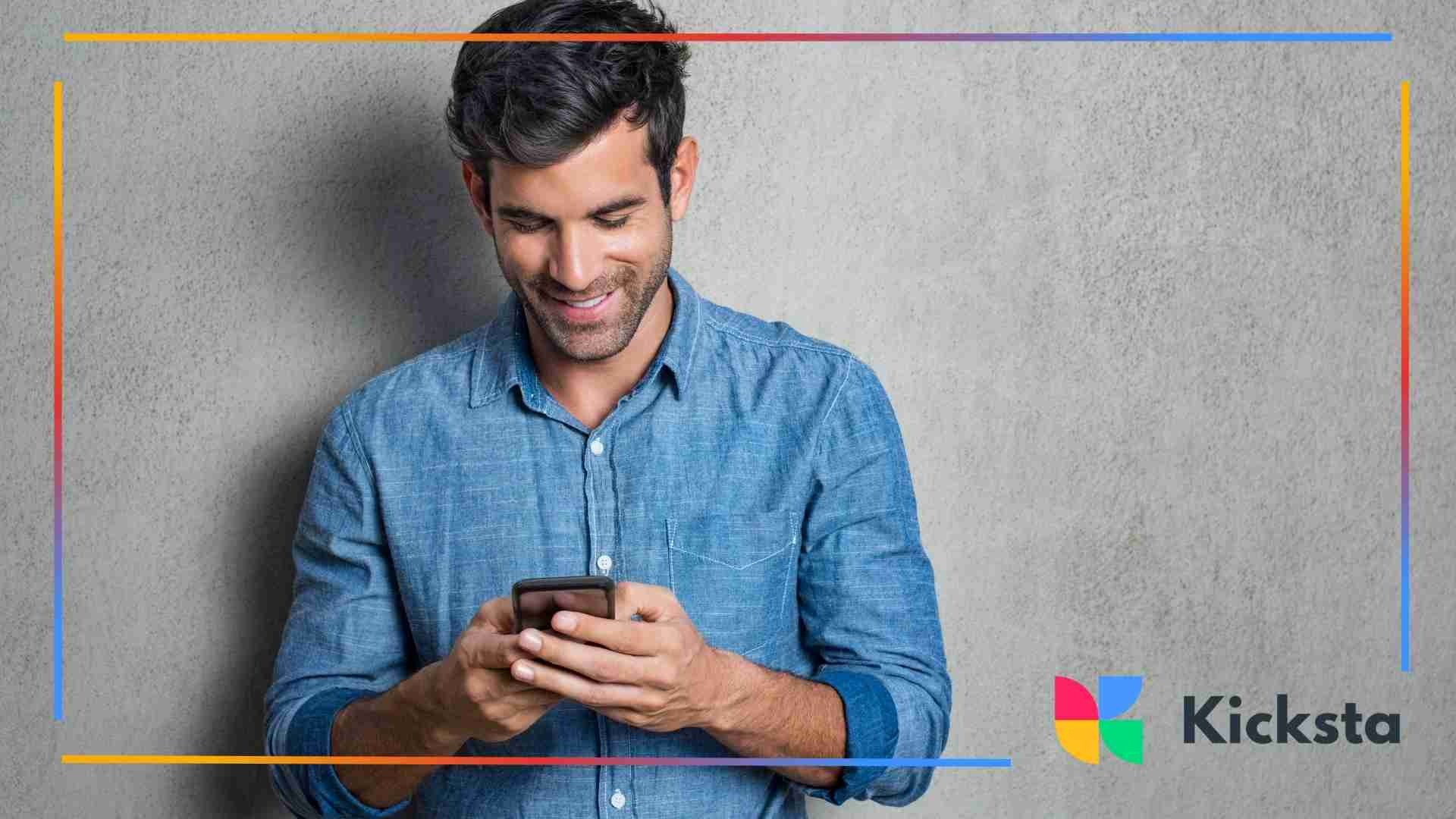 A man in a denim shirt stands against a gray wall while smiling at his smartphone.