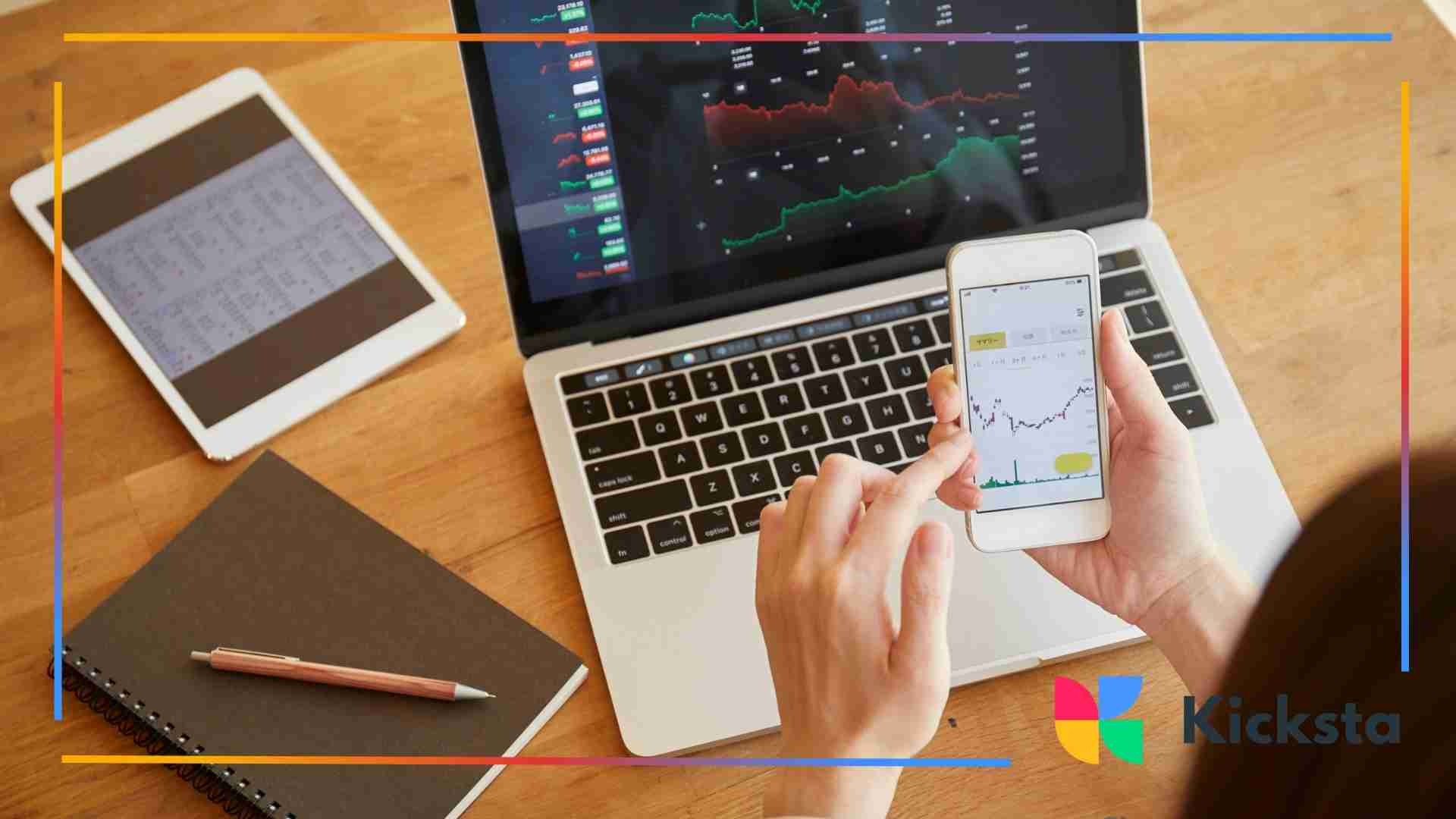 A person holding a smartphone in front of an open laptop displaying charts and graphs, with a tablet and notebook on the desk.