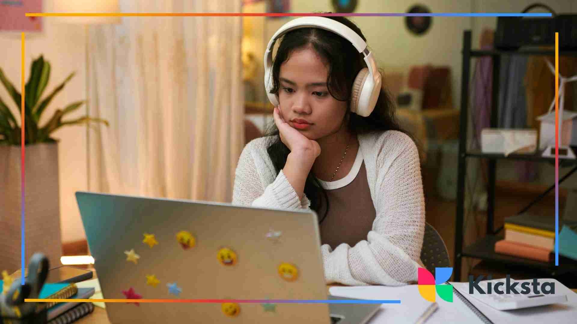 A young woman wearing large white headphones, resting her chin on her hand while looking thoughtfully at her laptop covered in emoji stickers.