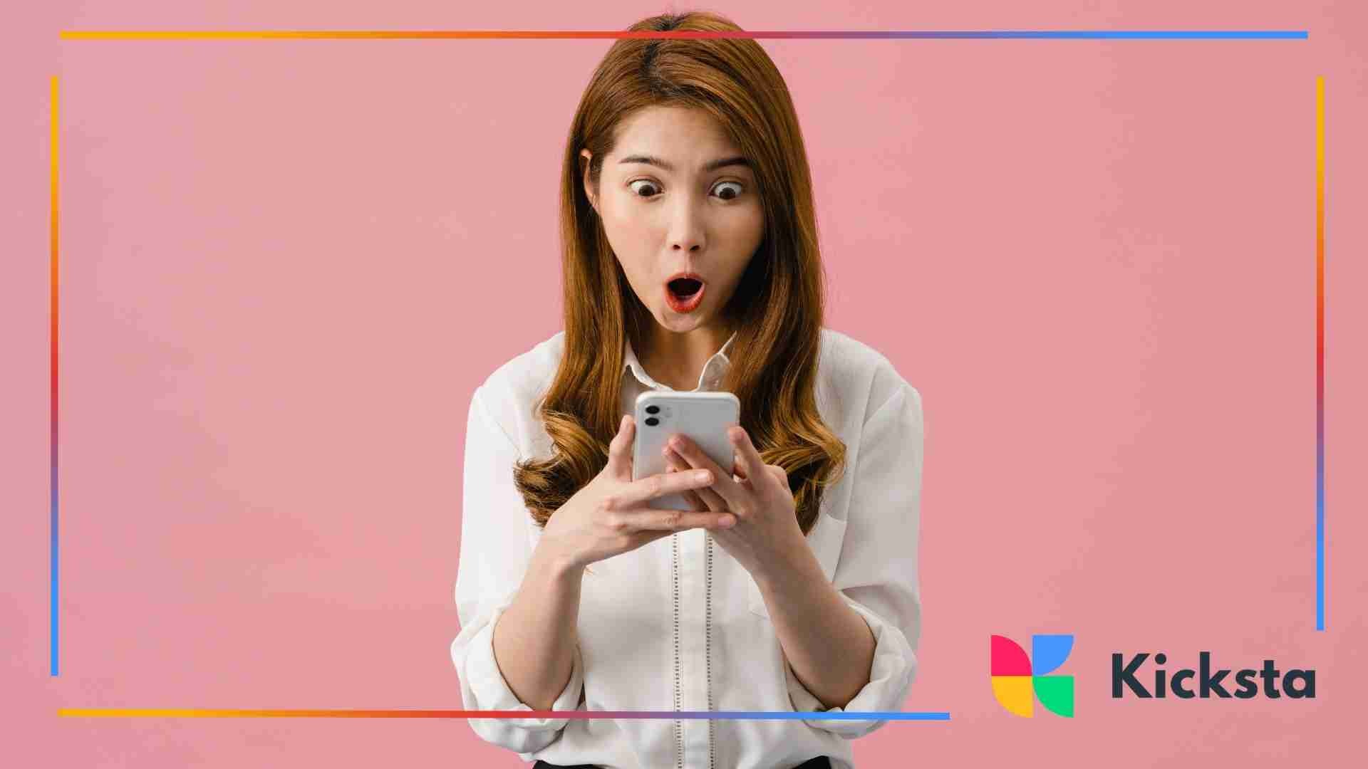 Woman with long hair in a white blouse looking at her phone with wide eyes and an expression of shock or surprise against a pink background.