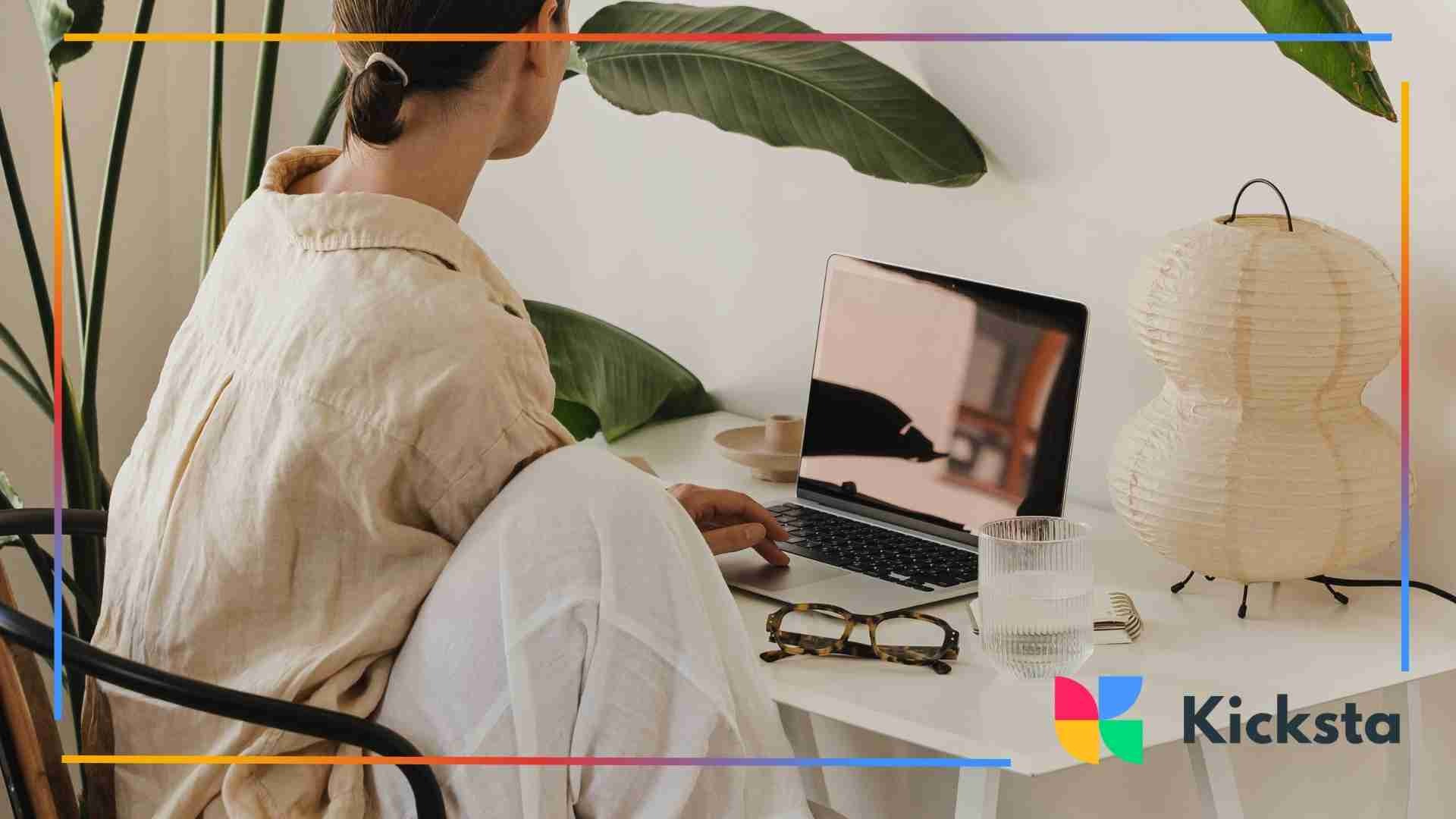 A woman sitting at a minimalist home workspace with a laptop, plants, glasses, and soft neutral décor.