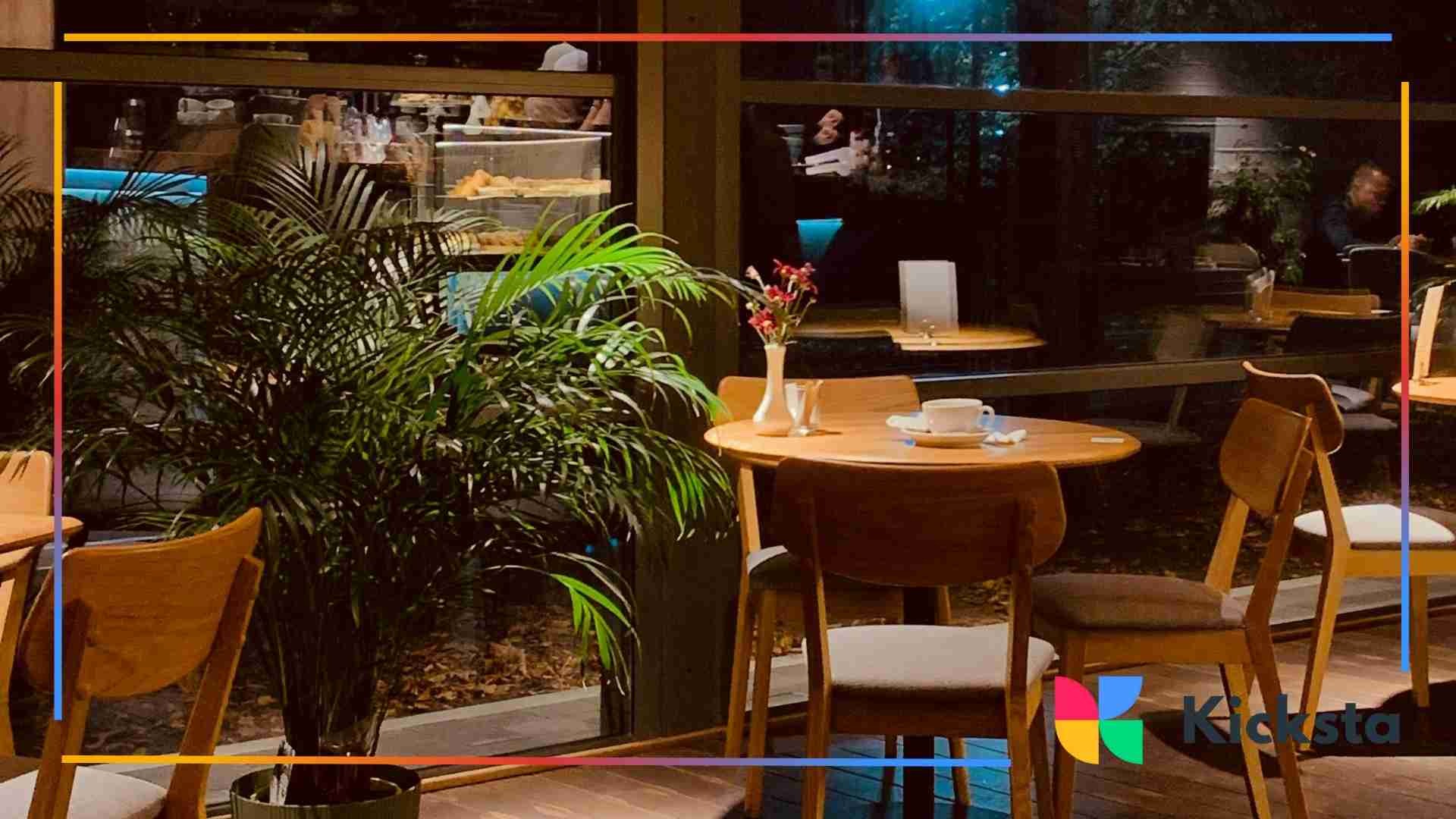 A warm and inviting café interior featuring wooden tables, chairs, indoor plants, and soft ambient lighting at night.