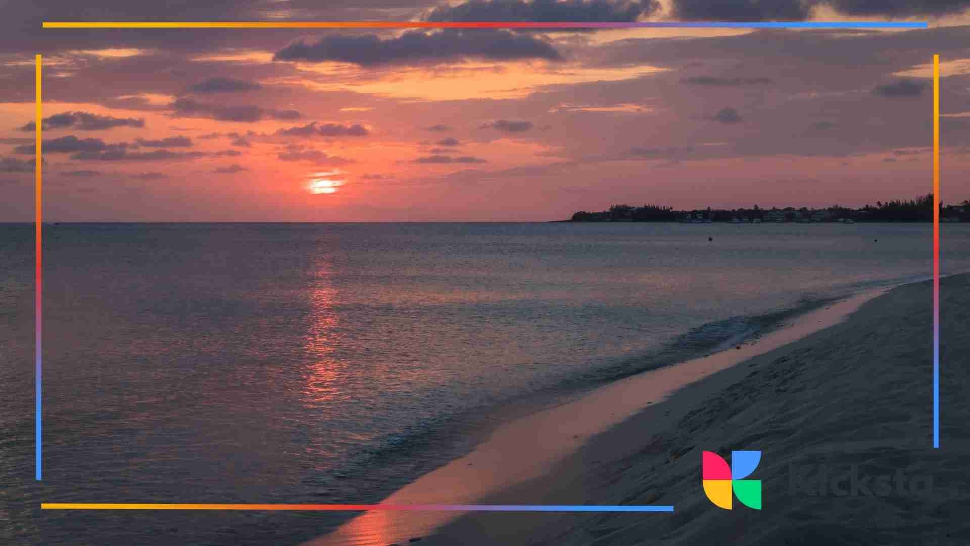 A serene beach at sunset with pink and orange skies reflecting over calm ocean waves and soft sand in the foreground.
