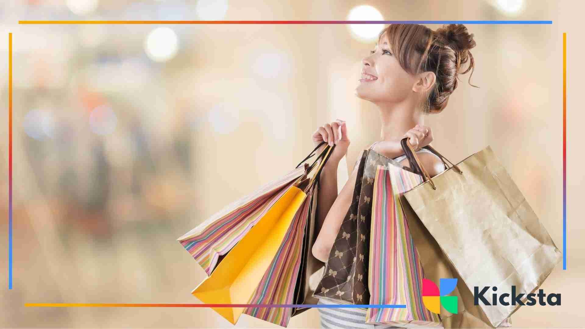 A smiling woman holding multiple colorful shopping bags in both hands inside a brightly lit mall.