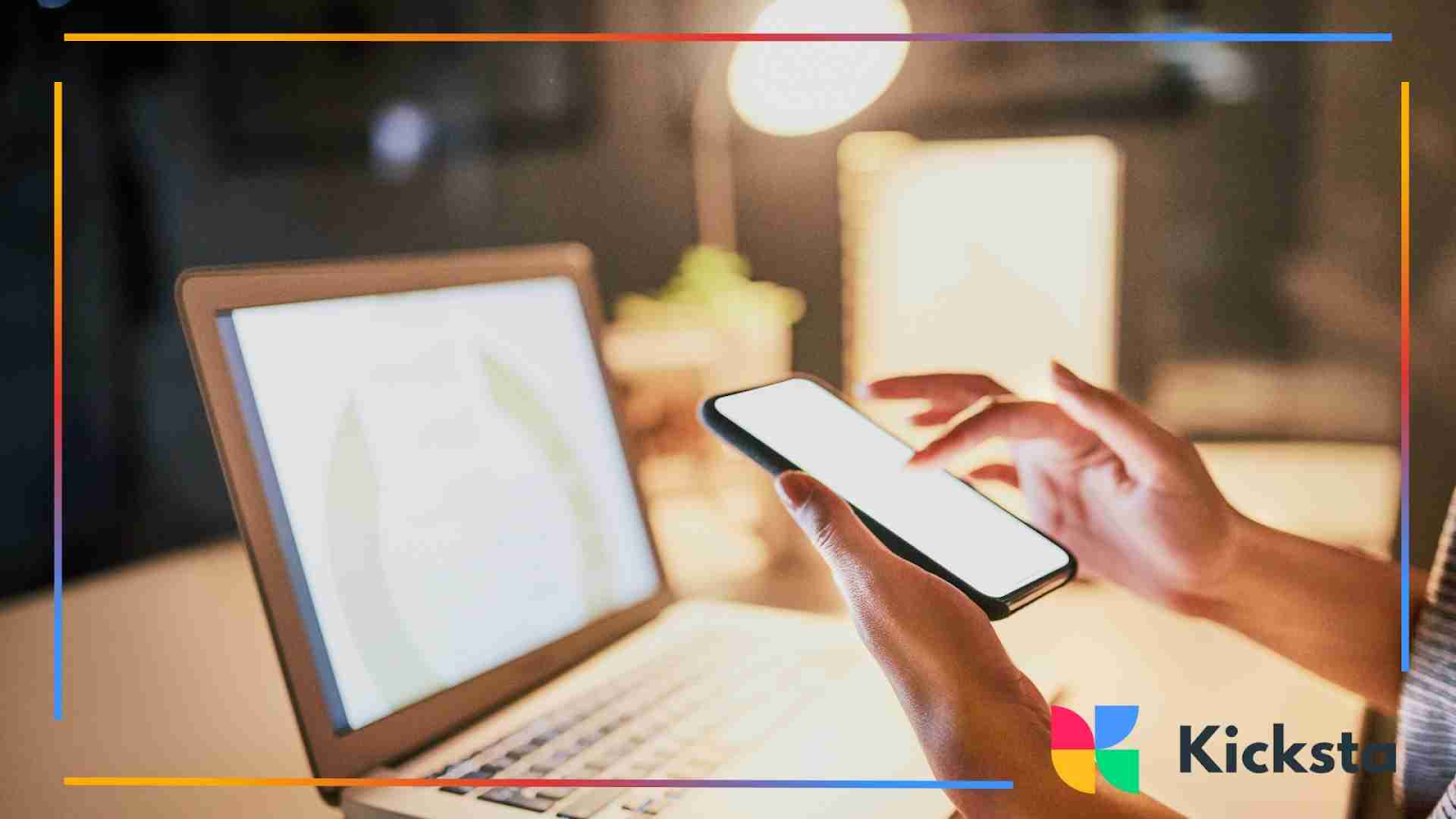 A person holds a smartphone in one hand while working on a laptop under warm desk lighting.