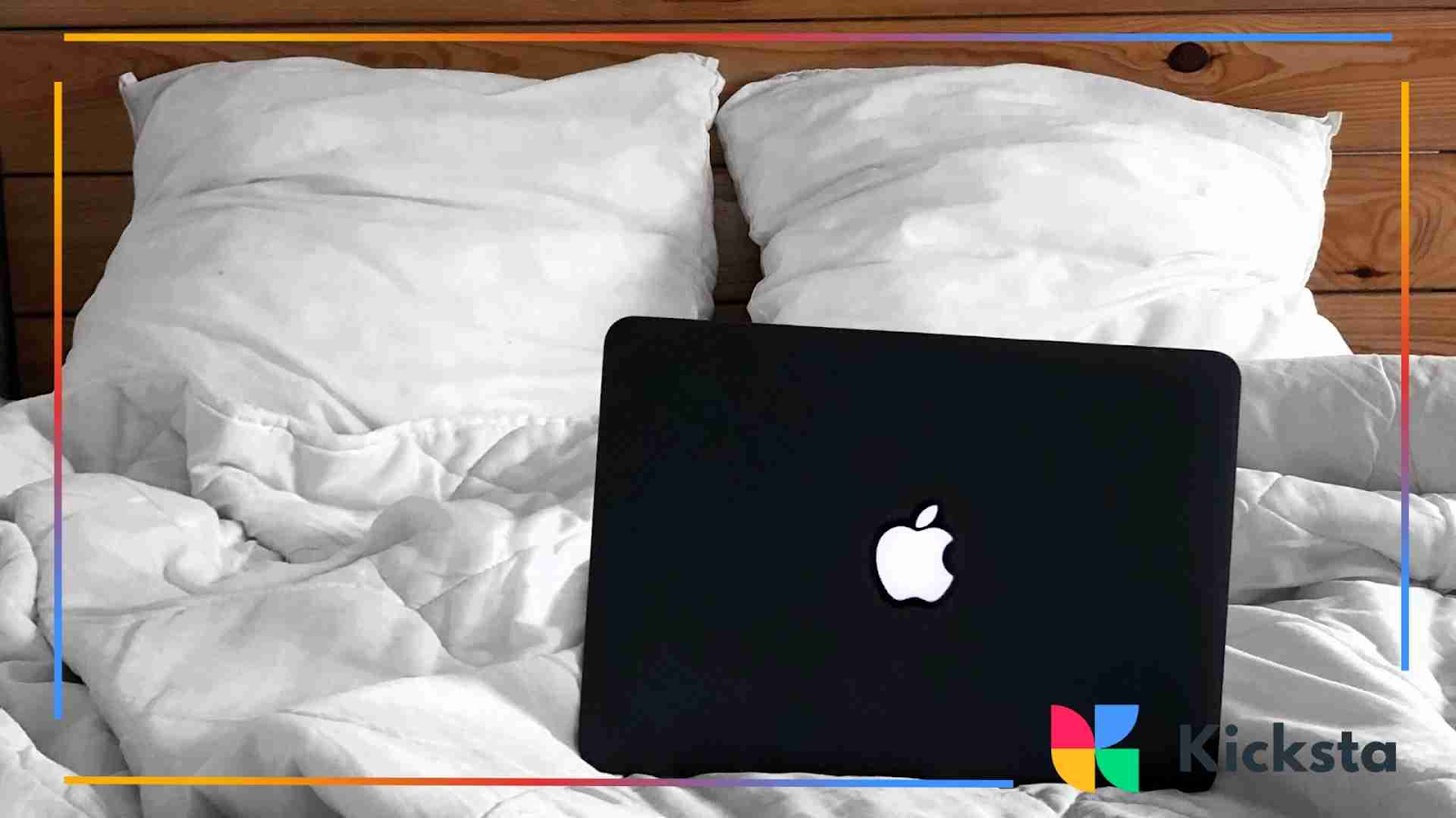 A black laptop with an illuminated logo sits on a white, unmade bed with pillows behind it.