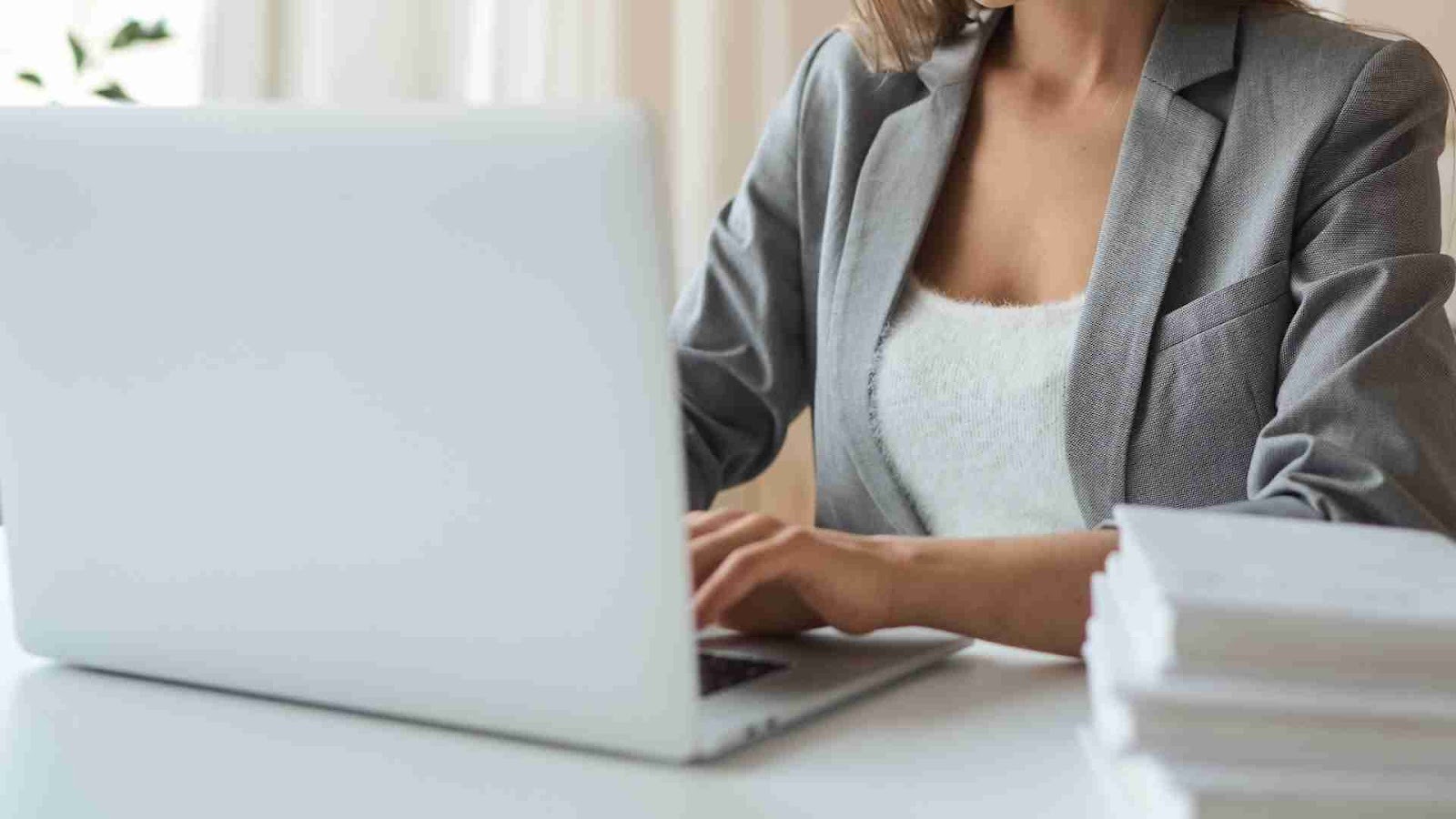 Close-up of a person in a gray blazer typing on a silver laptop at a clean white desk.