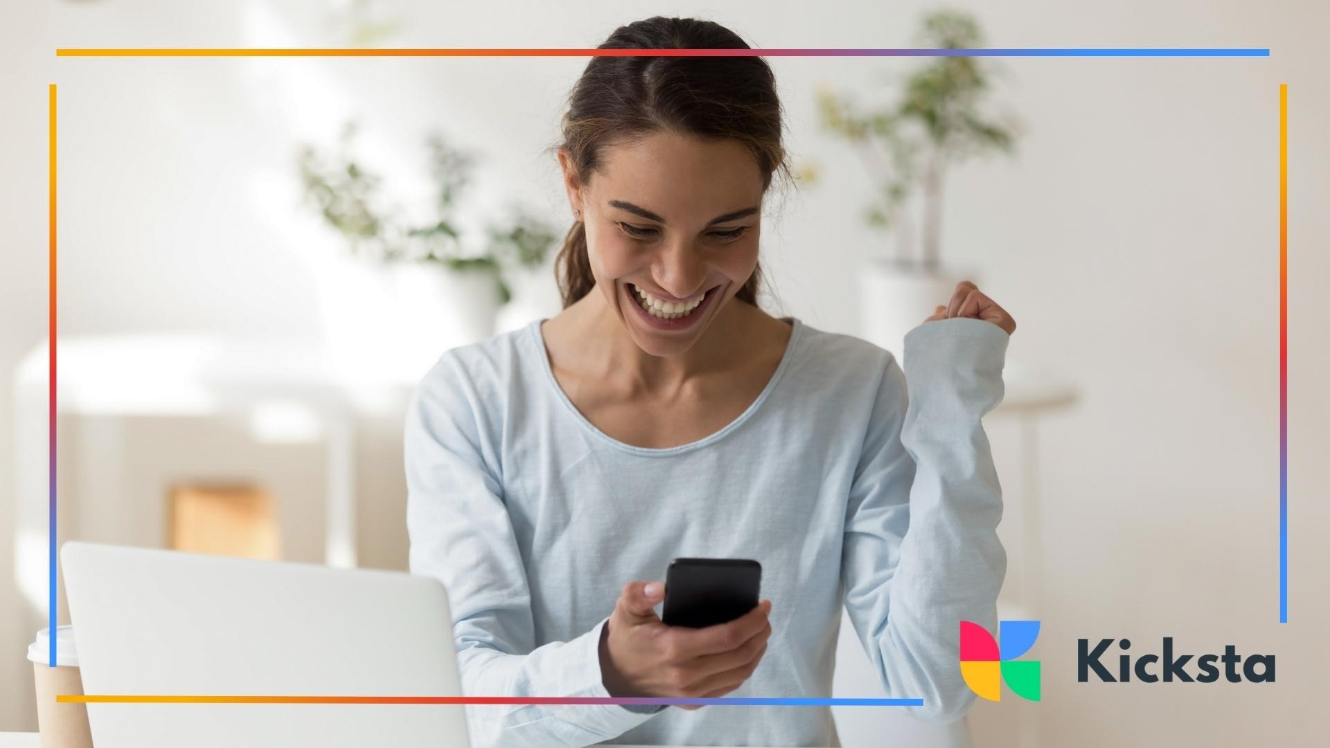A smiling woman celebrating while looking at her phone, sitting at a desk with a laptop in a bright, minimalist room.