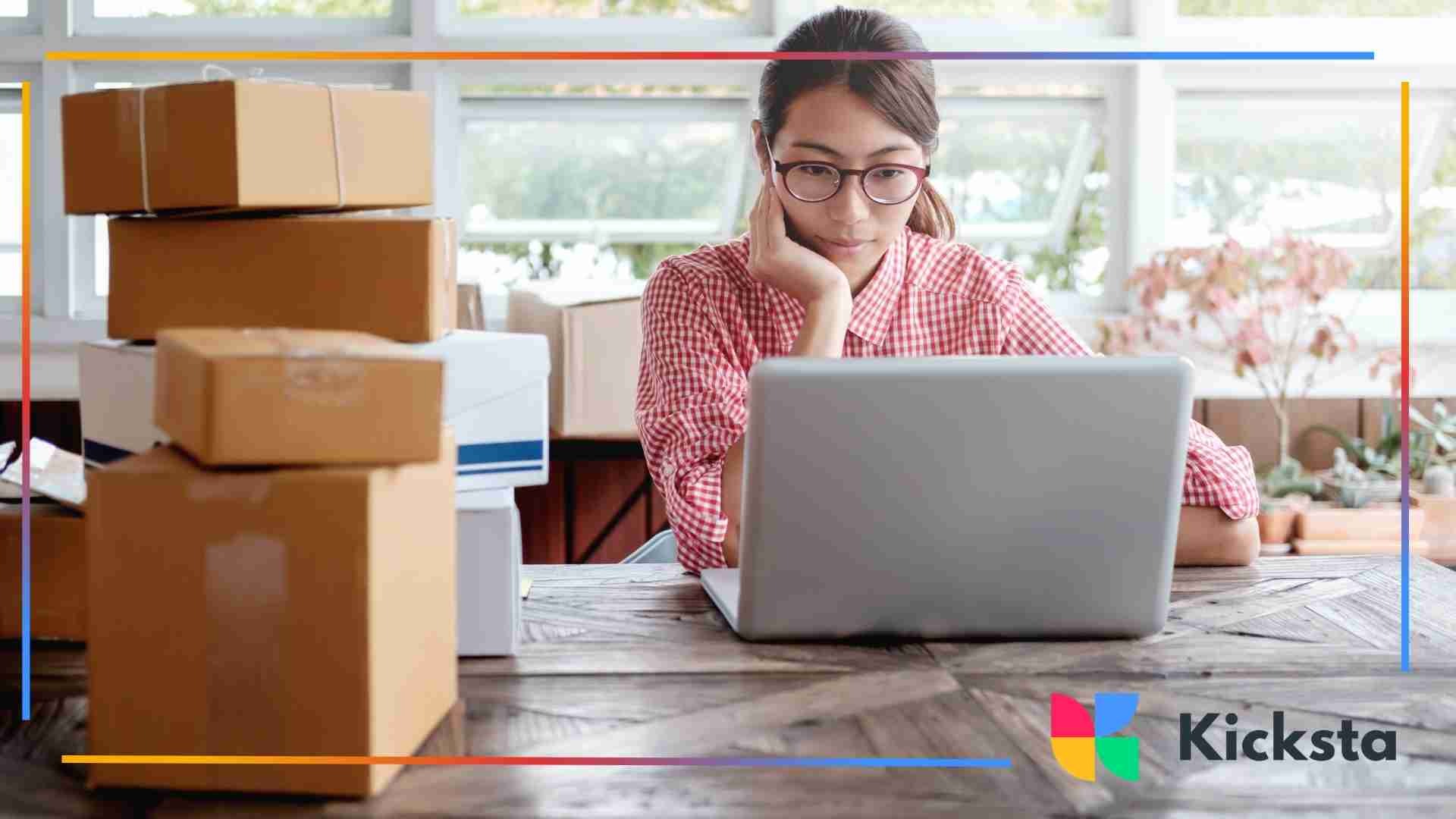 Small business owner surrounded by shipping boxes while working on a laptop at a wooden table.