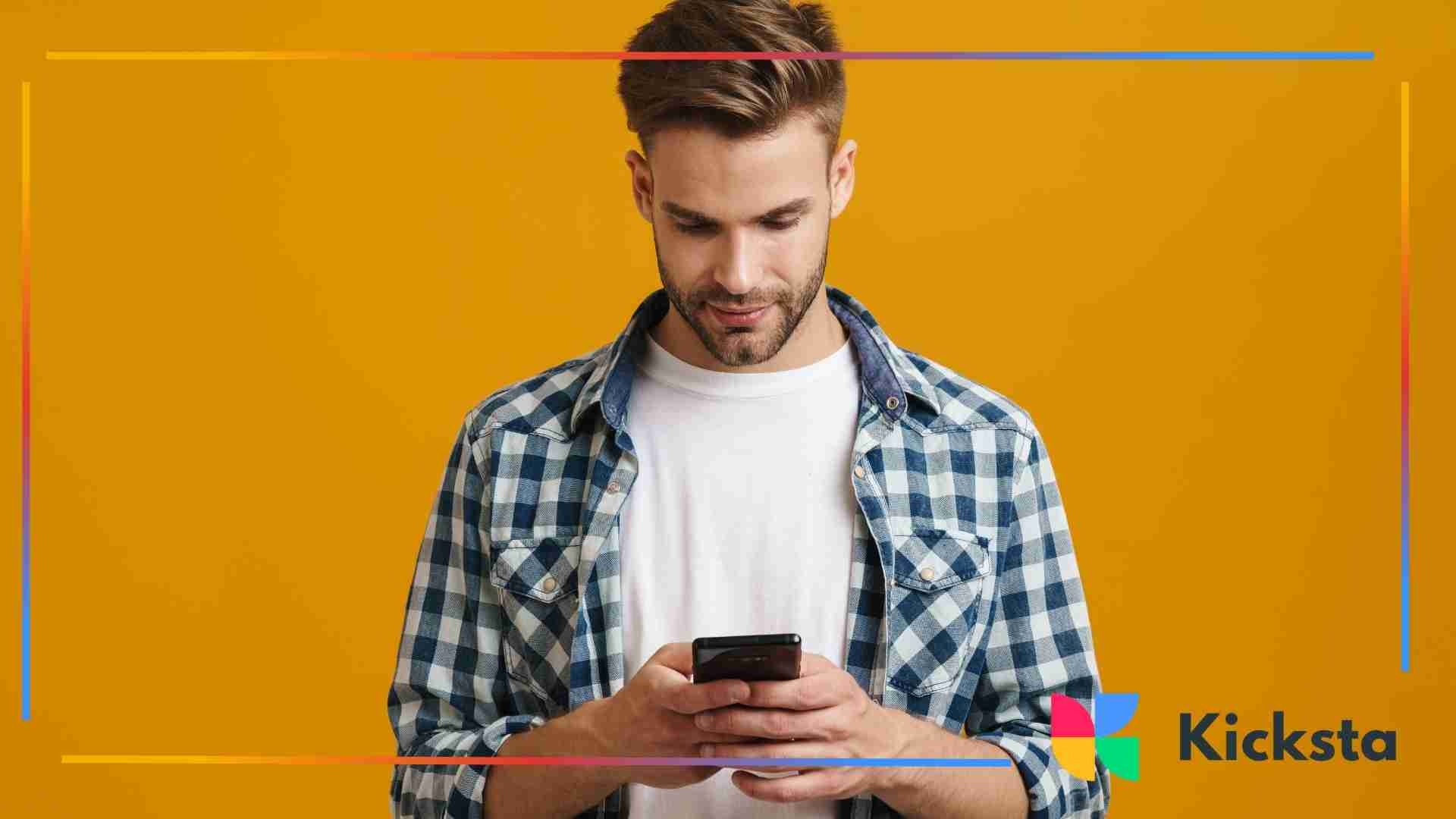 Man in a checkered shirt standing against a bright yellow background while looking down at his phone.