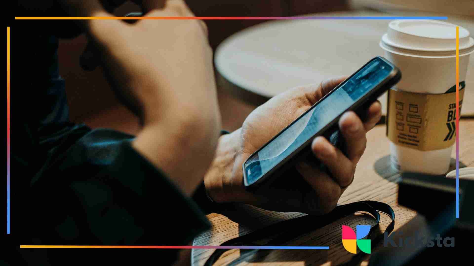 Close-up of a person holding a smartphone with one hand while sitting at a table with a coffee cup in the background.