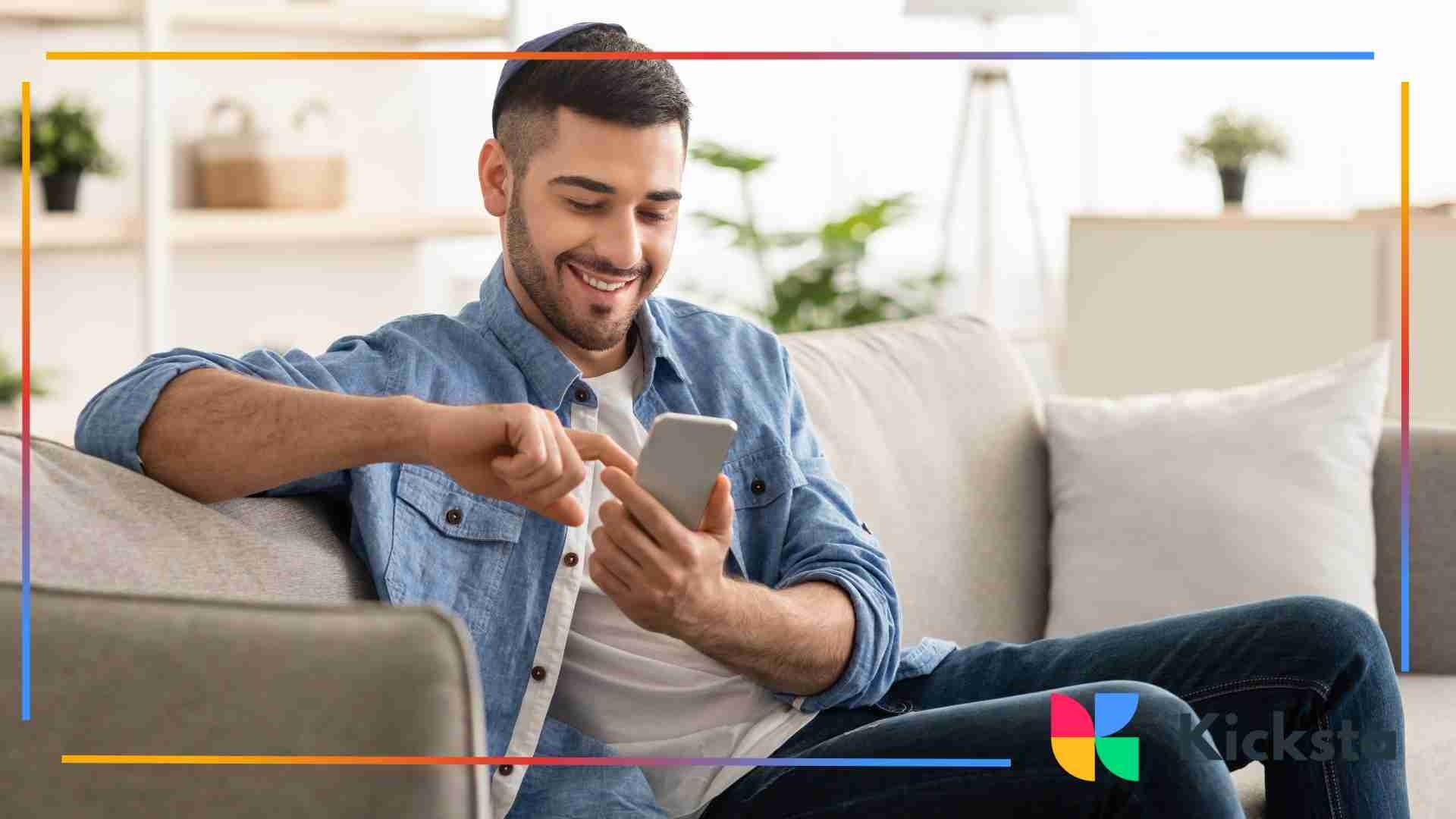 Smiling man relaxing on a couch while using his smartphone in a bright, cozy living room.