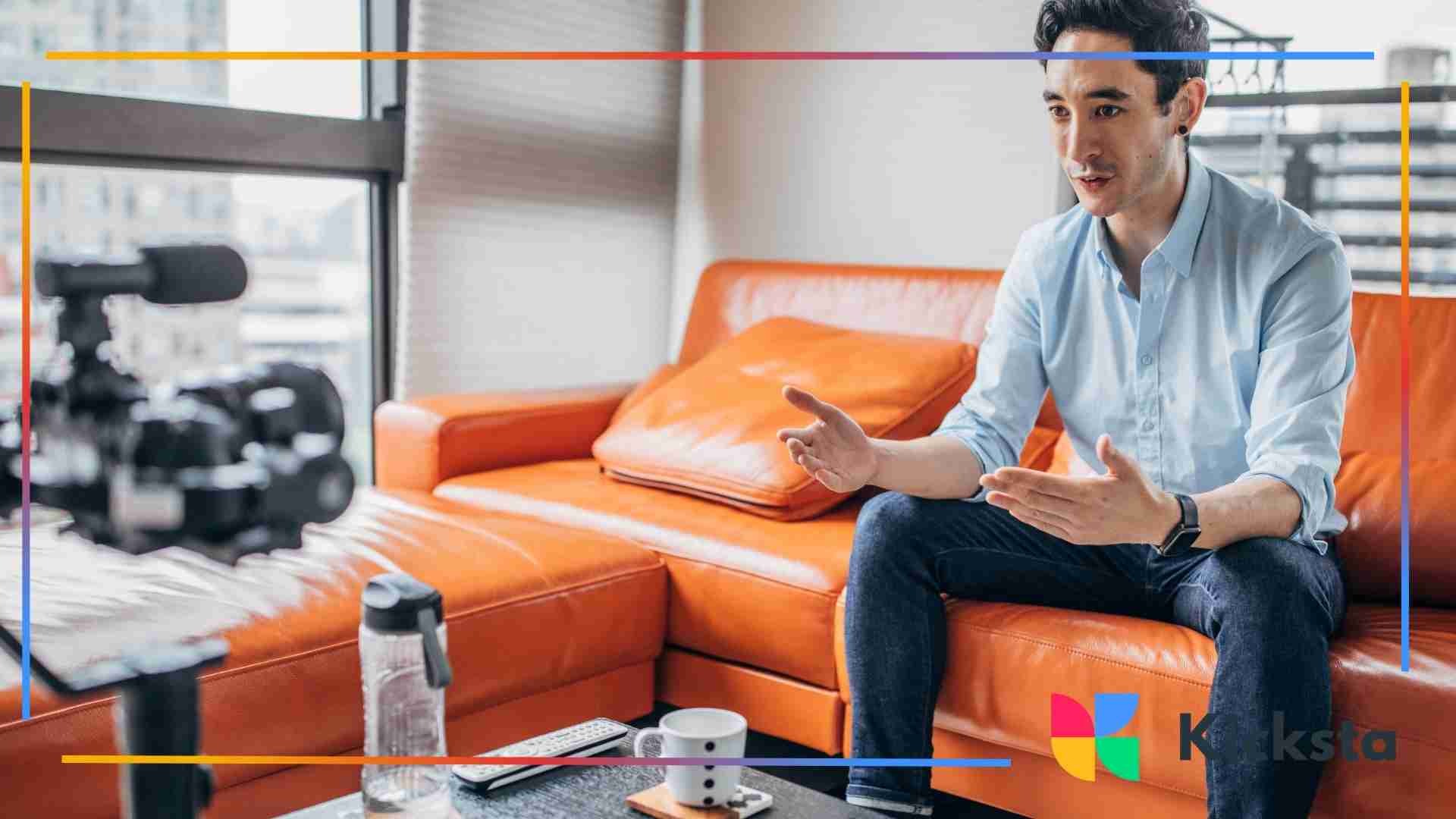 A man sitting on an orange sofa, speaking expressively toward a camera set up on a tripod in a bright living room.