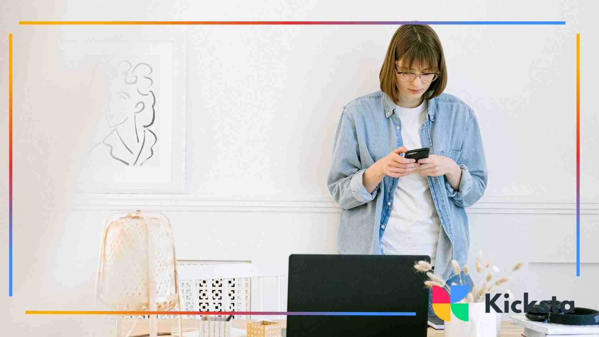 A woman wearing glasses and a denim shirt stands in a bright, minimal home office while looking down at her smartphone.