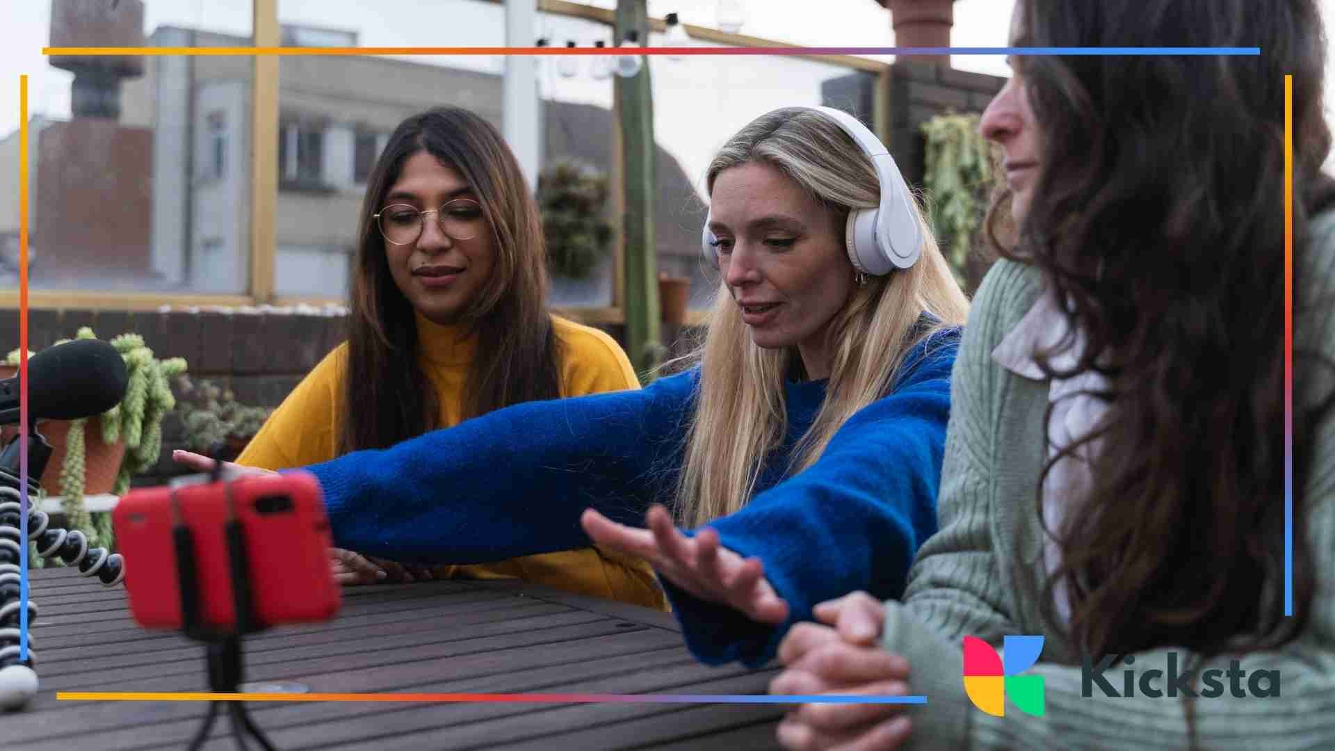 Three women sit outdoors at a table, with one woman extending her arm toward a smartphone on a tripod as they record content together.