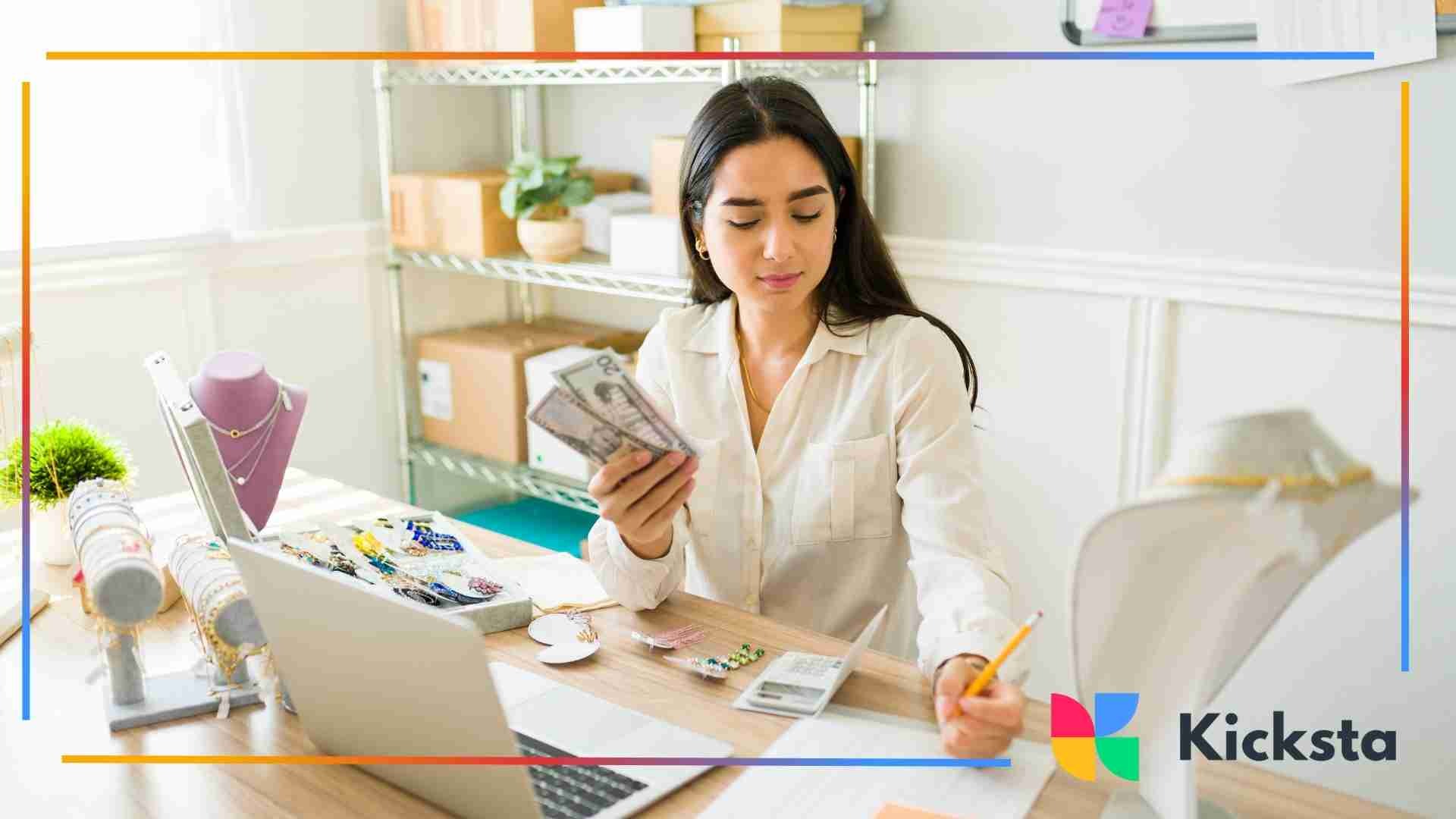 Woman at a workspace counting cash while managing jewelry products, paperwork, and a laptop.