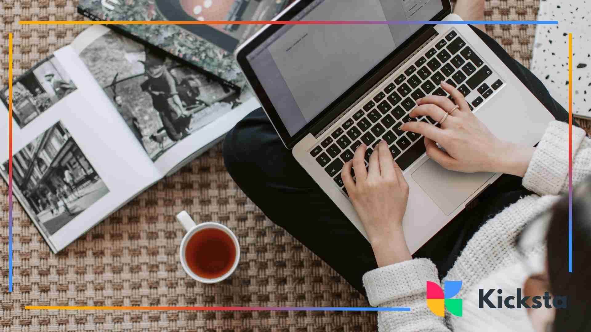 Person typing on a laptop while sitting on the floor next to an open photo book and a cup of tea.