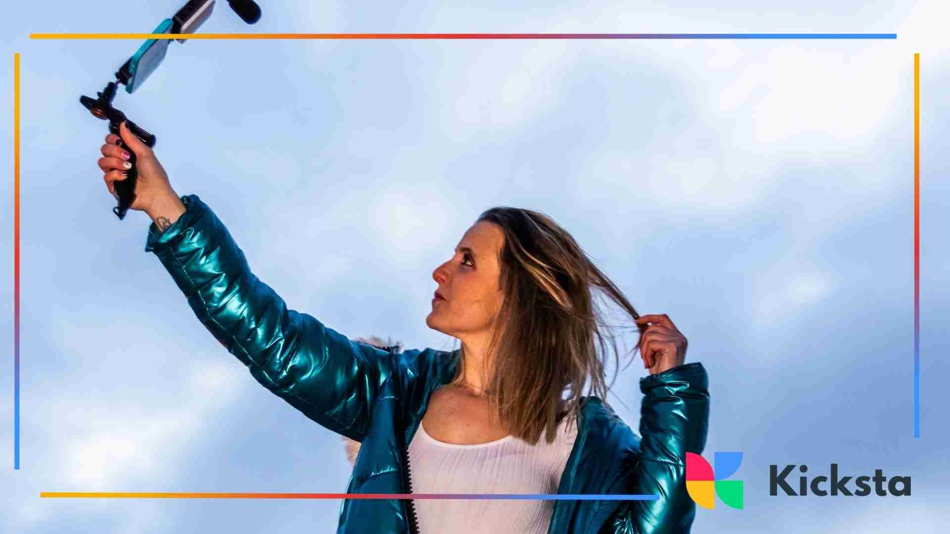 Woman outdoors holding a selfie stick with a smartphone, adjusting her hair while filming.