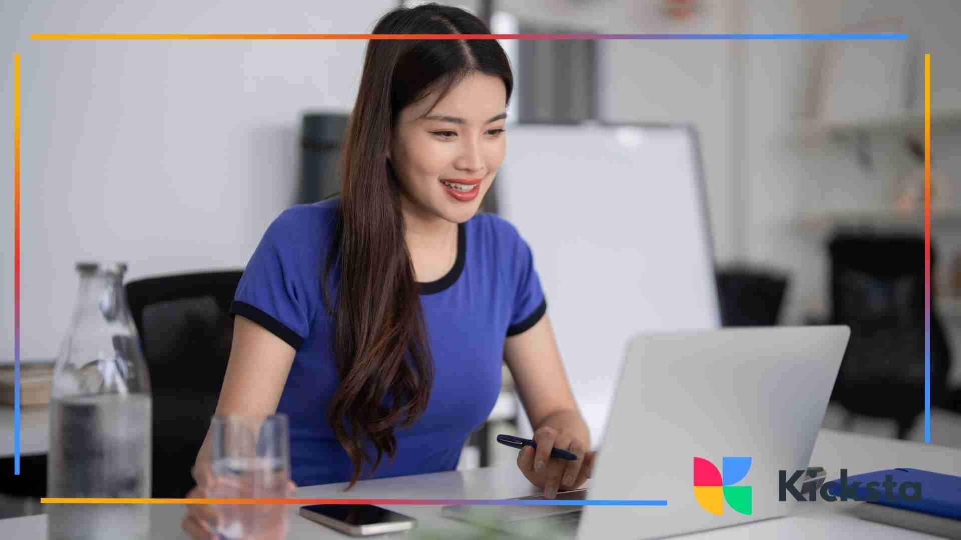 Woman smiling while working on a laptop at a desk, holding a pen with a water bottle and glass nearby.