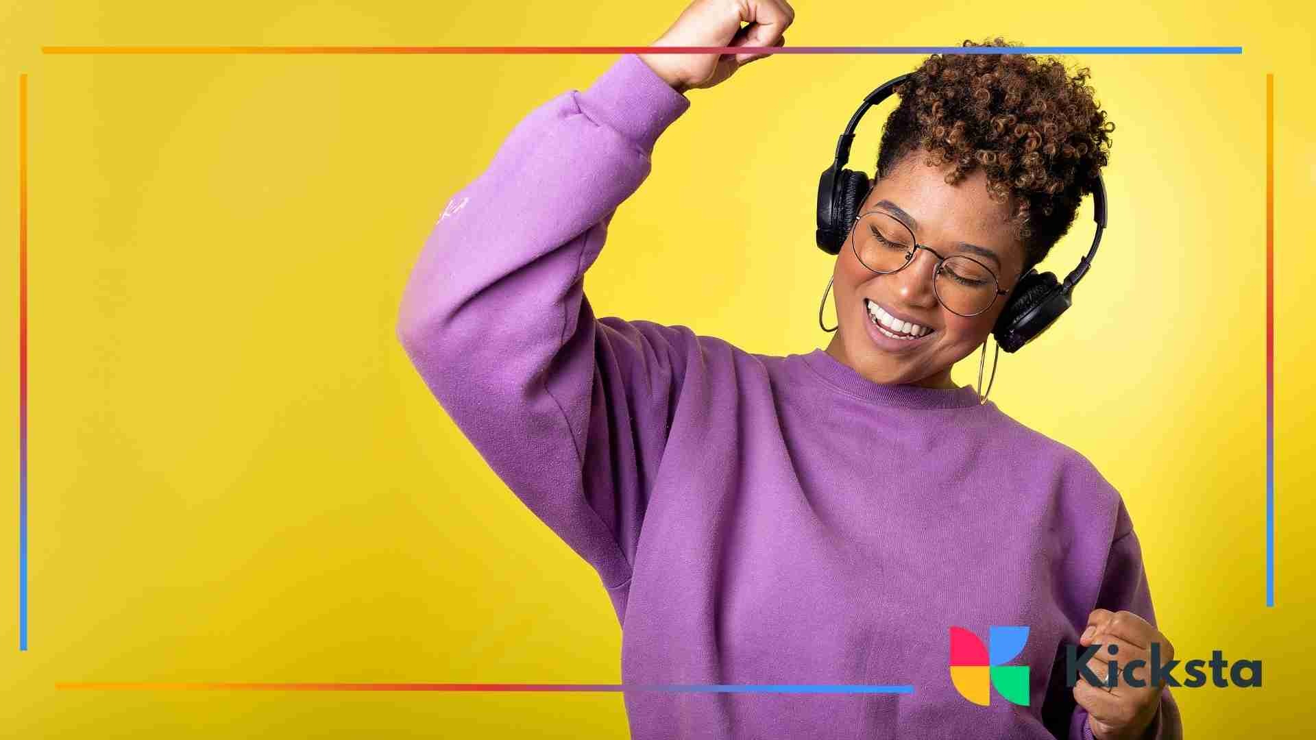 Woman in a purple sweater smiling and dancing while listening to music on headphones against a bright yellow background.