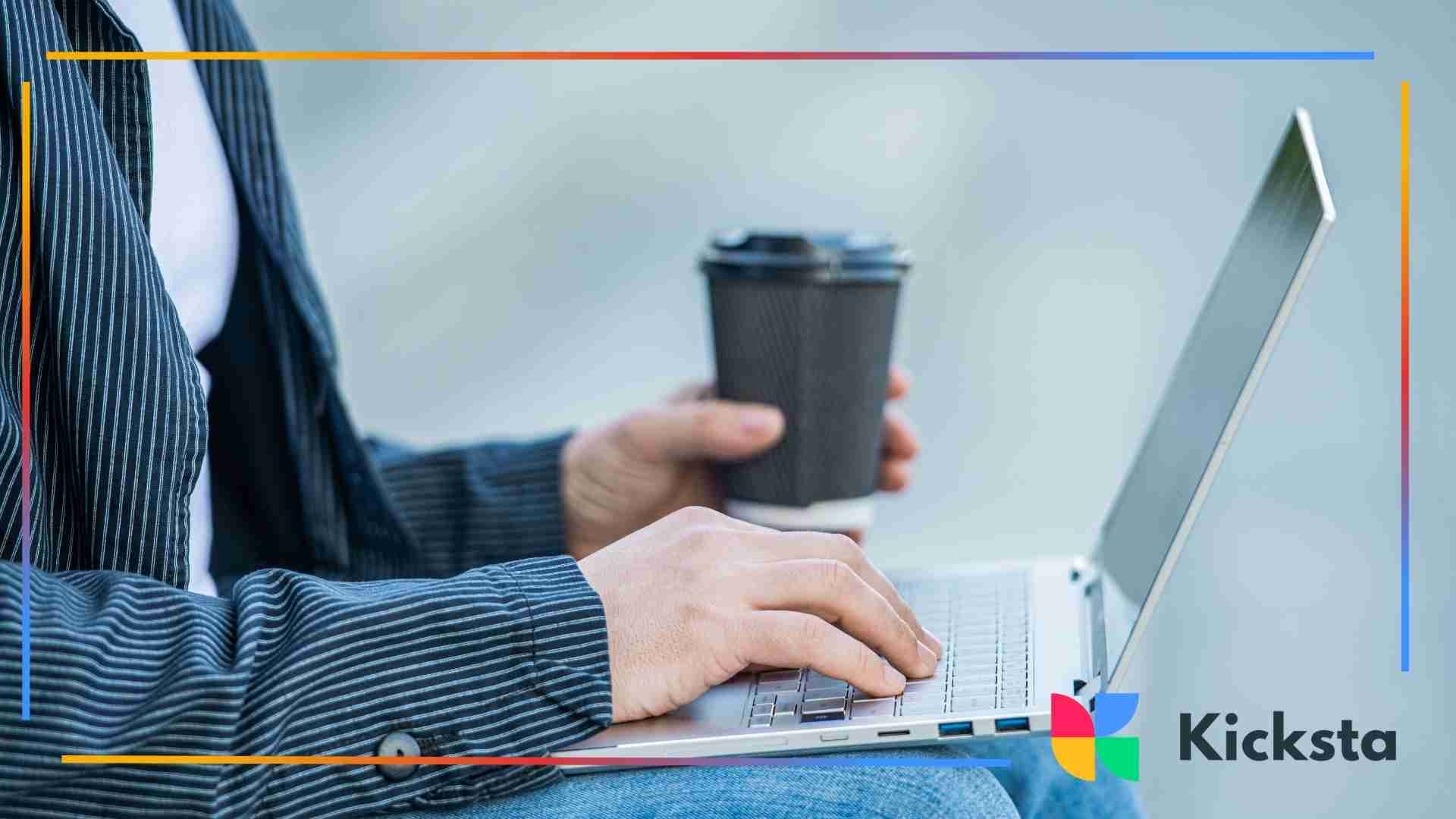 Person holding a coffee cup while typing on a laptop outdoors, wearing a striped shirt and jeans.
