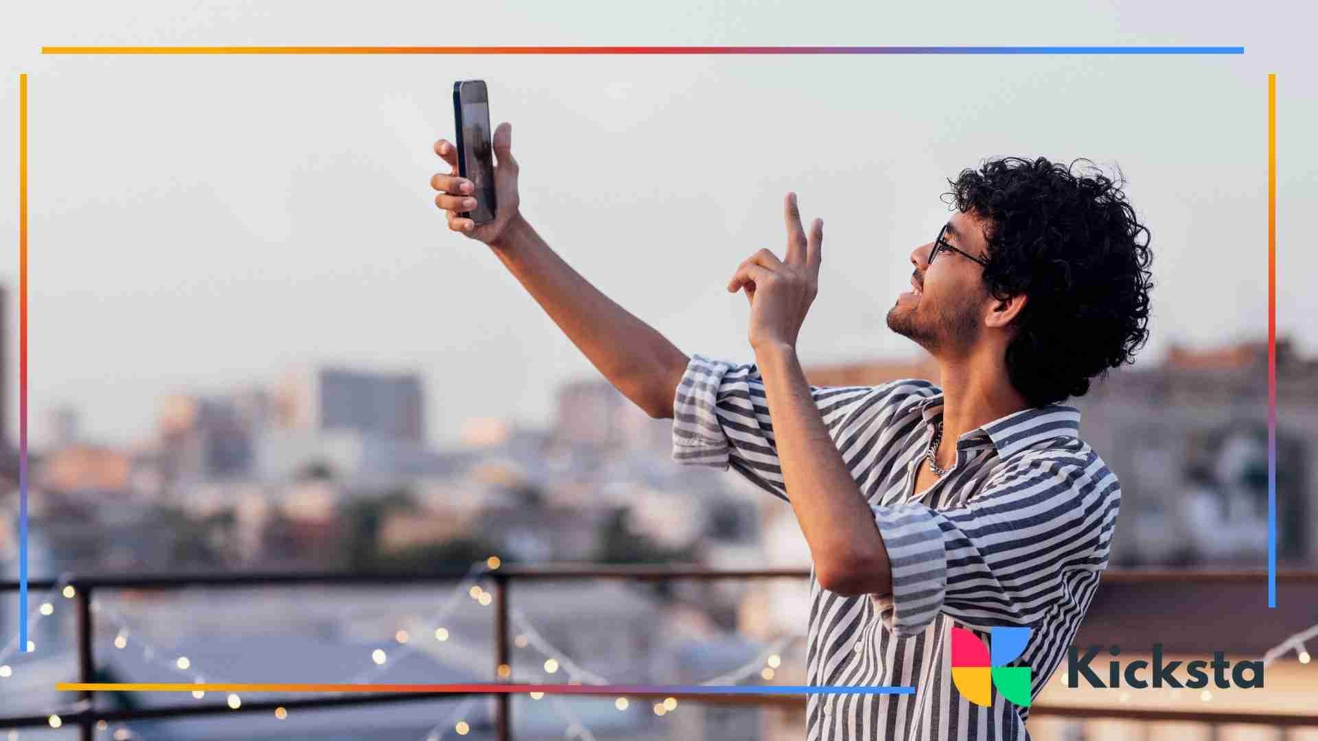 Man taking a selfie video on a rooftop at sunset, smiling and gesturing while holding his phone up.