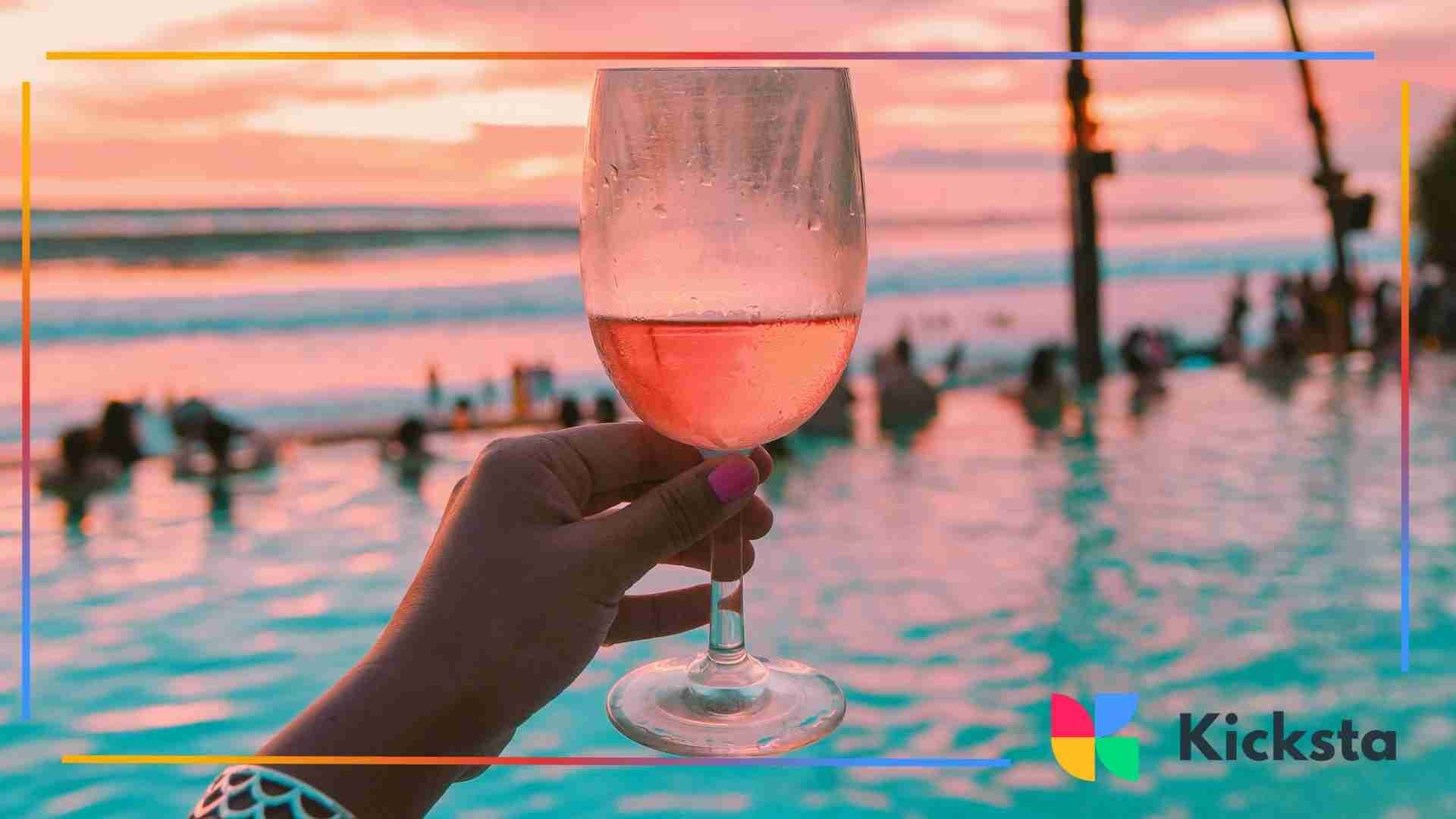 A hand holding a glass of rosé wine by a pool at sunset with people swimming in the background.