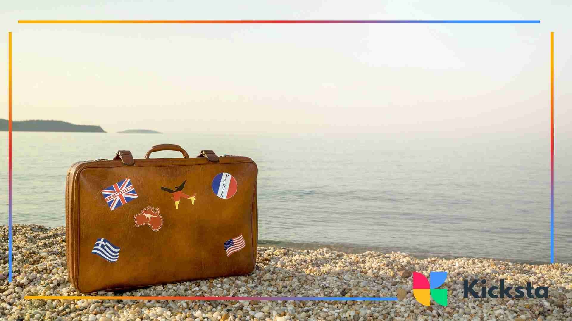 A vintage brown suitcase decorated with travel stickers sits on a rocky beach with calm ocean water in the background.